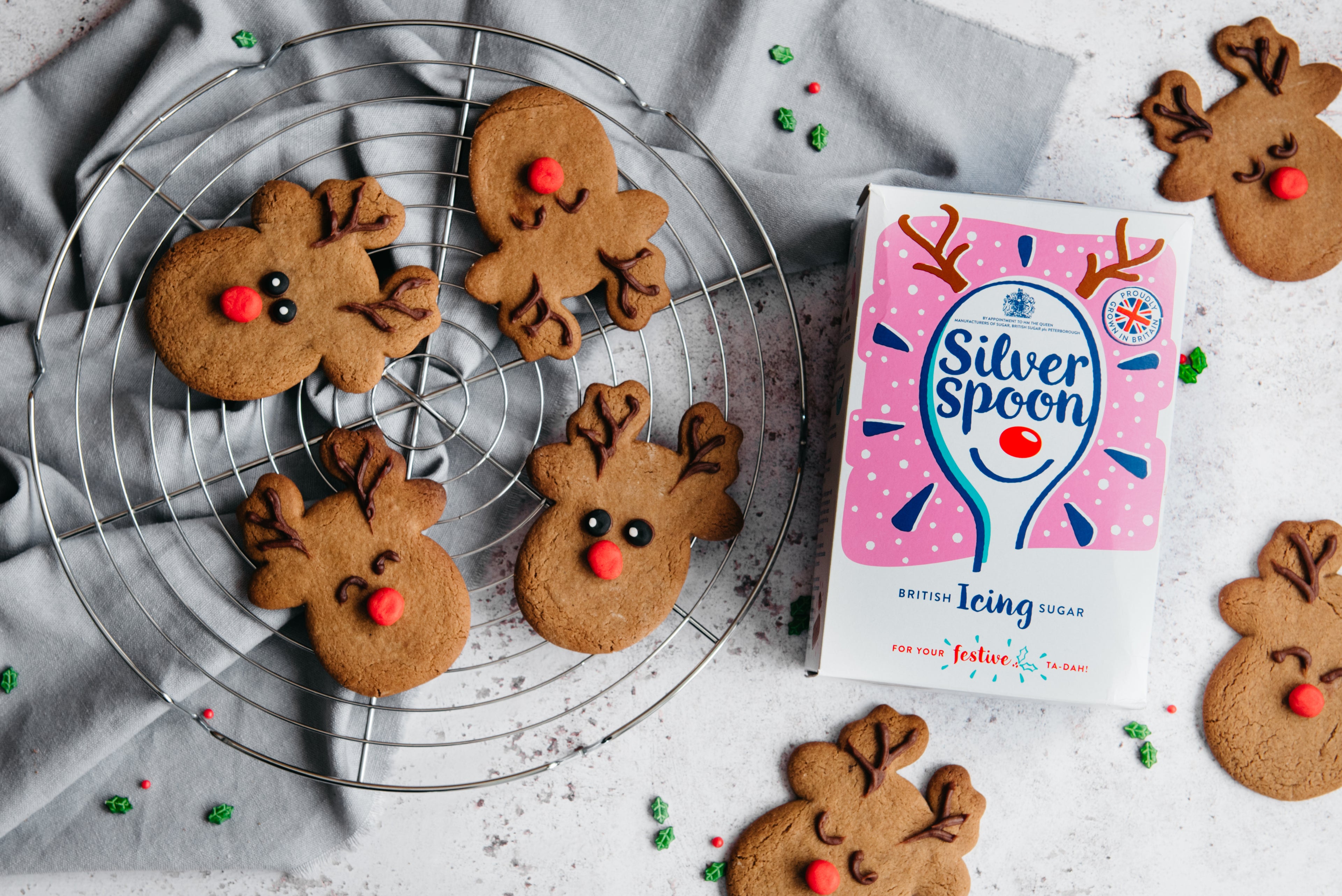 Reindeer Cookies on a wire cooling rack, hand decorated with reindeer faces, next to a box of Silver Spoon icing sugar