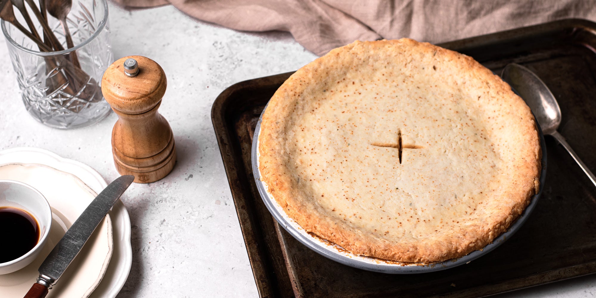 Suet Crusted Beef & Onion Pie on a baking tray, next to a pepper mill and a set of plates with a knife ready to serve