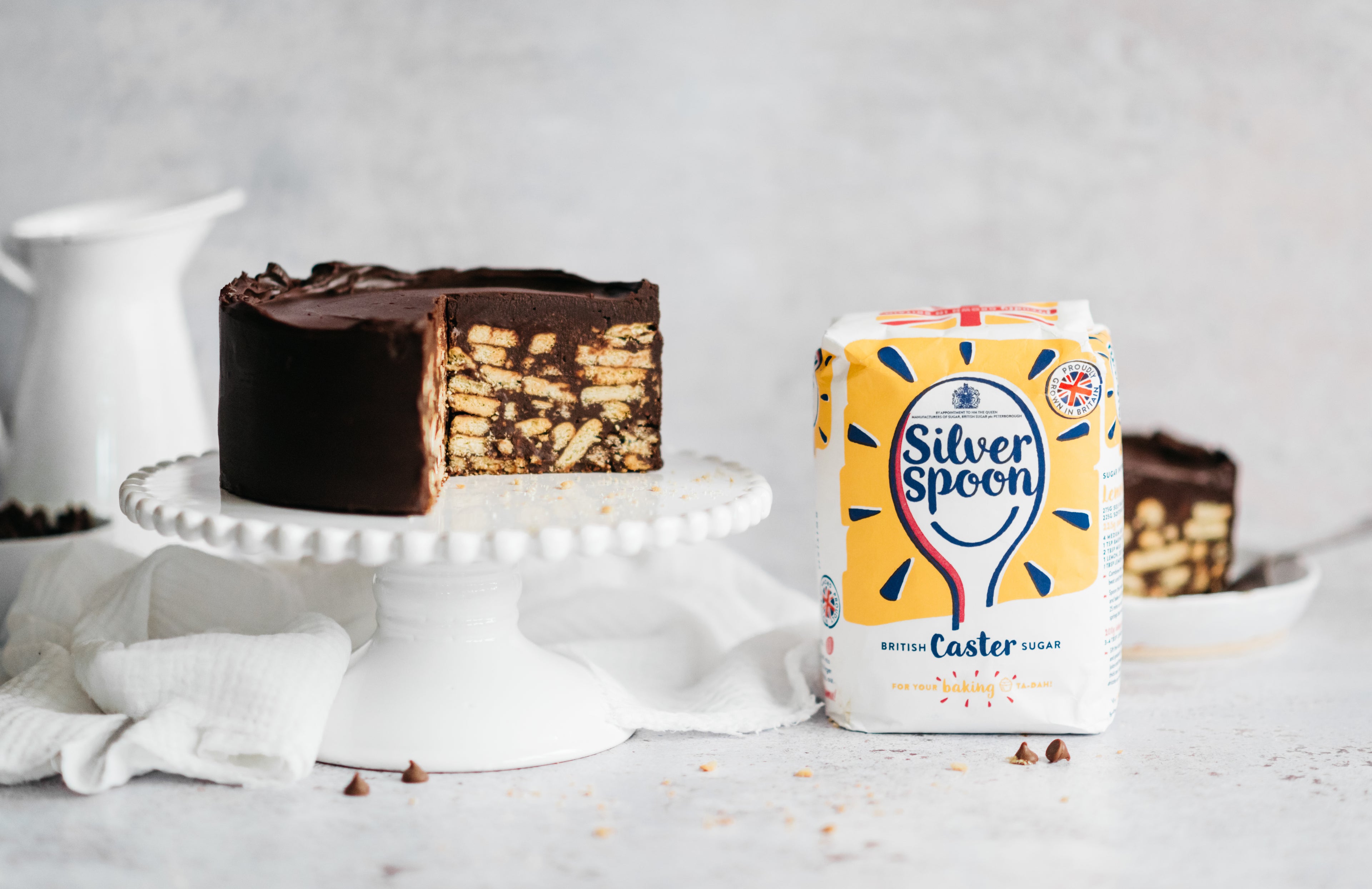 Chocolate Biscuit Cake served on a cake stand with a slice cut out showing the rich tea biscuit centre and chocolate filling. Next to a bag of Silver Spoon Caster Sugar