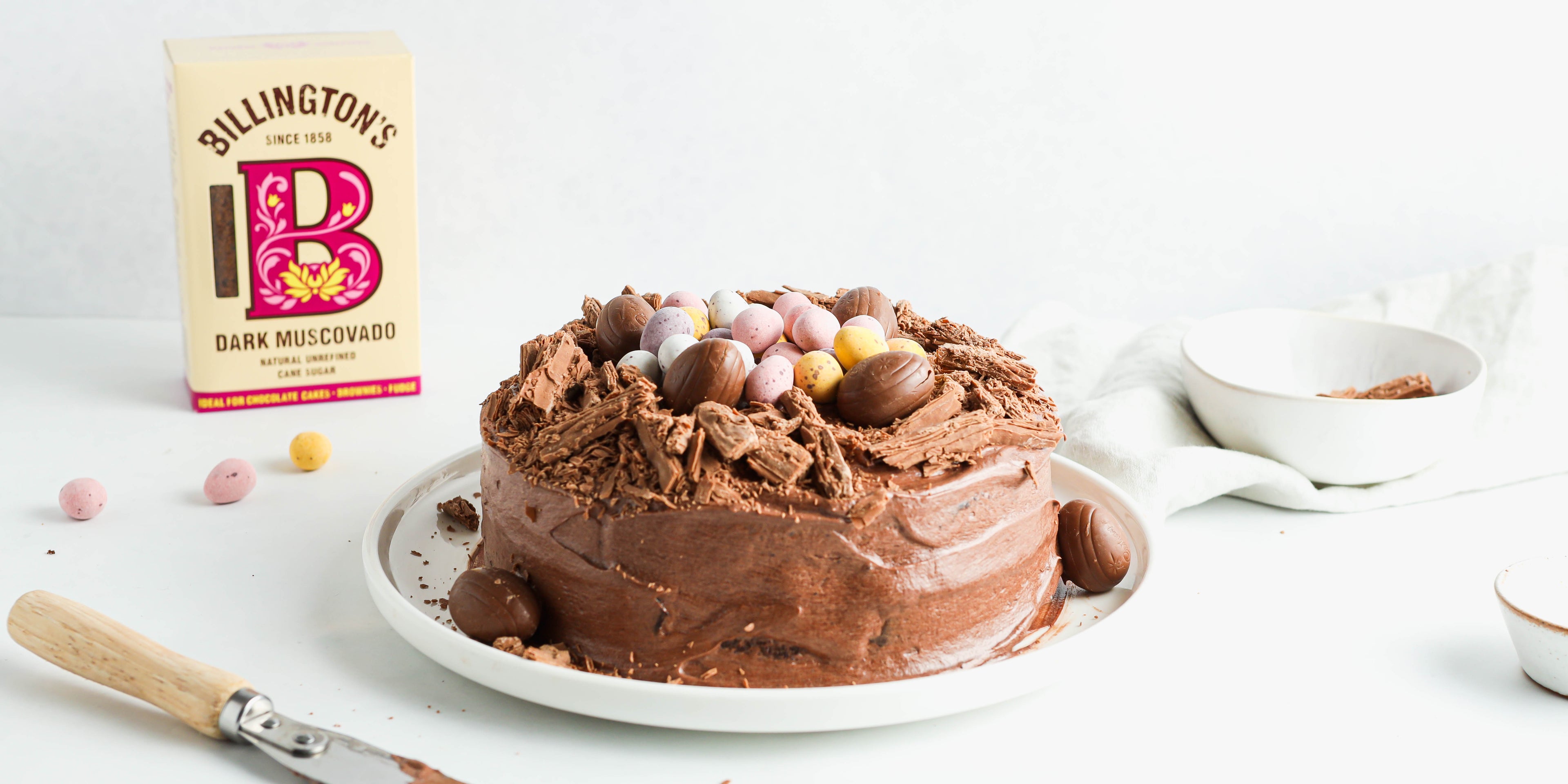Finished Chocolate Nest Cake topped with easter eggs, and crushed flake, in front of a box of Billington's Dark Muscovado sugar