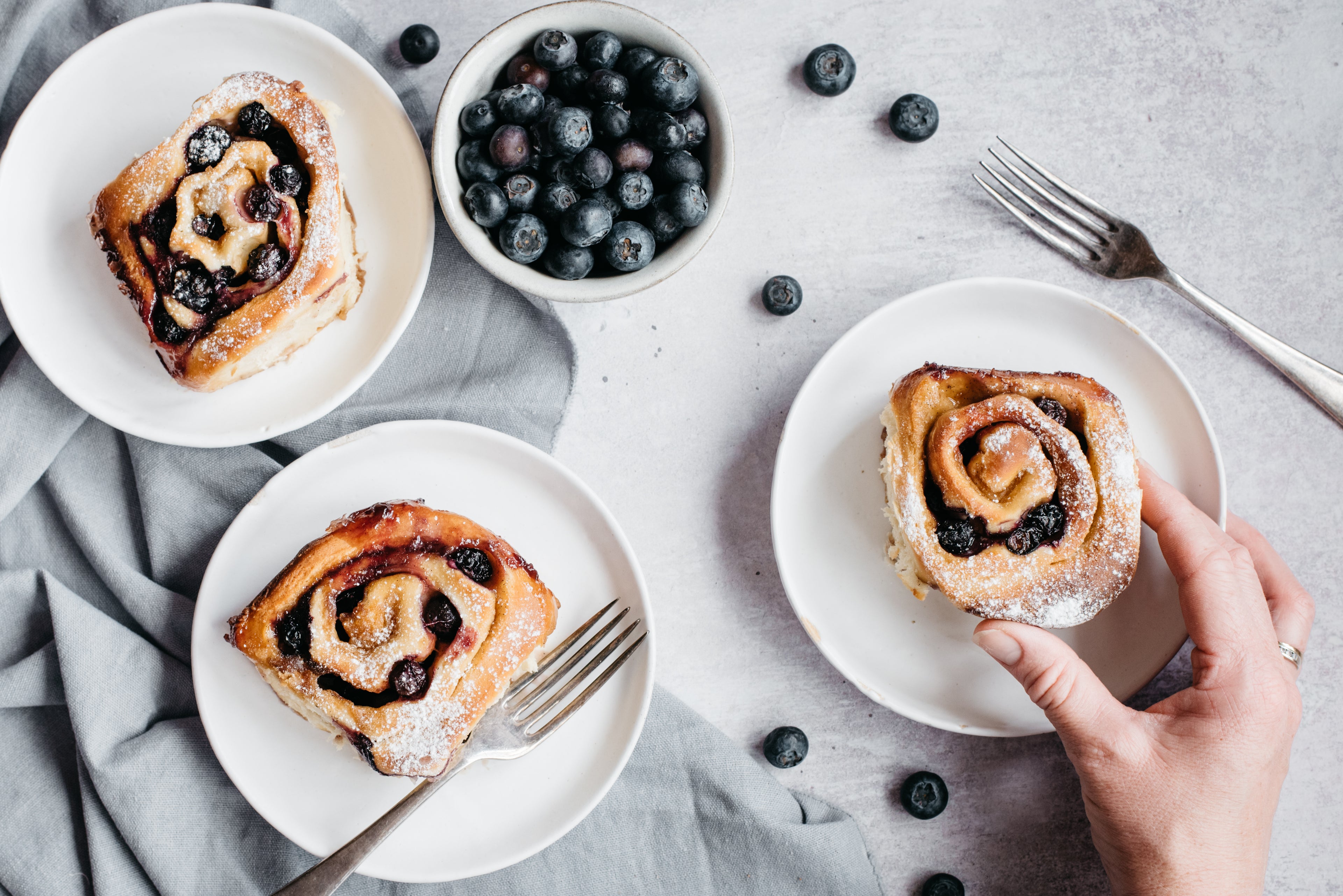 Three blueberry and vanilla rolls on plates, with forks and hand