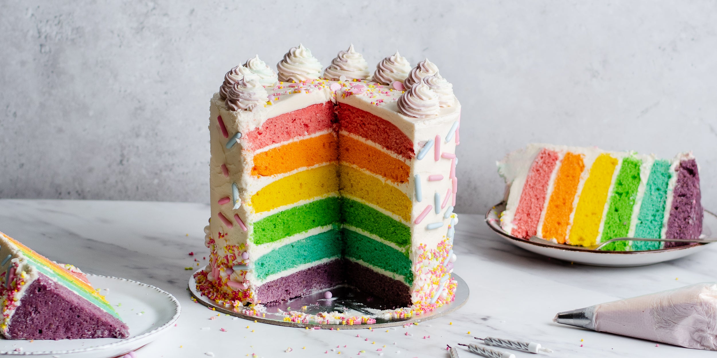 6 layer rainbow sponge cake cut open to show the layers. Two slices on white plates beside it. With piping bag and candles in forefront