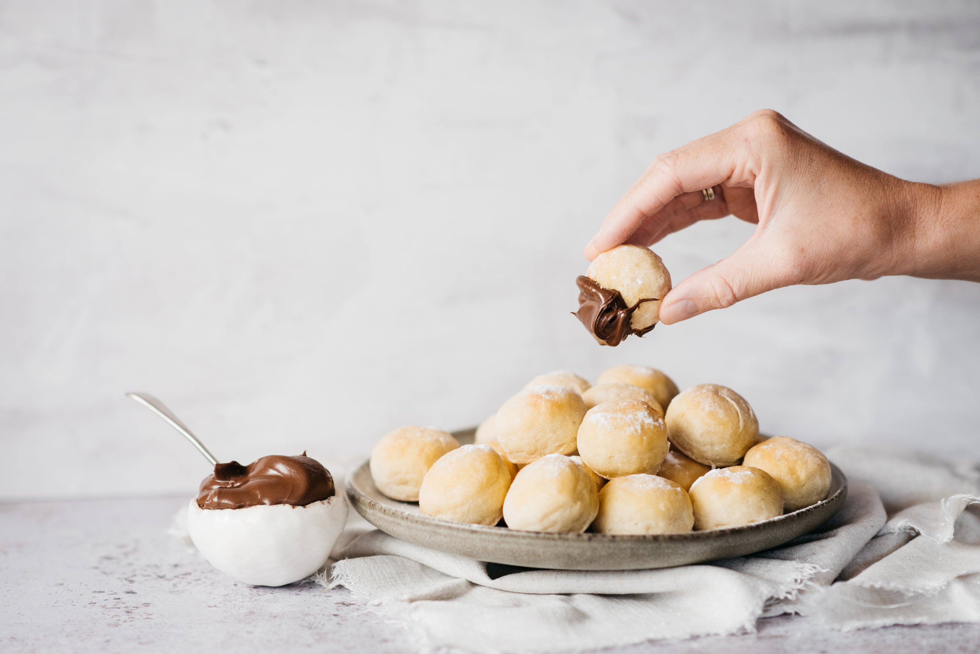 Plate of Chocolate Dough Balls with a hand holding a ball dipped in chocolate sauce. Next to a bowl of chocolate sauce, with a spoon