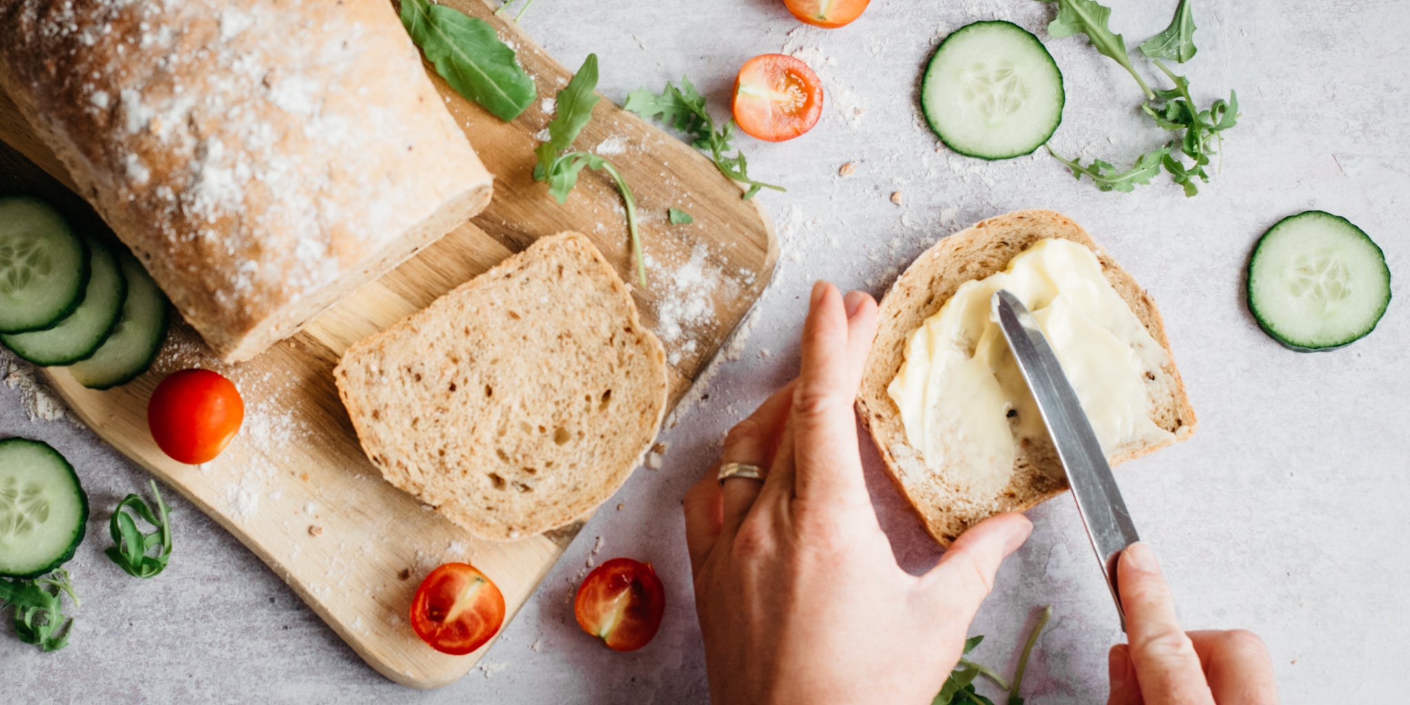 Top view of Farmhouse Country Grain Loaf sliced on a serving board. A hand is buttering a slice with a knife, surrounded by garnishes of salad