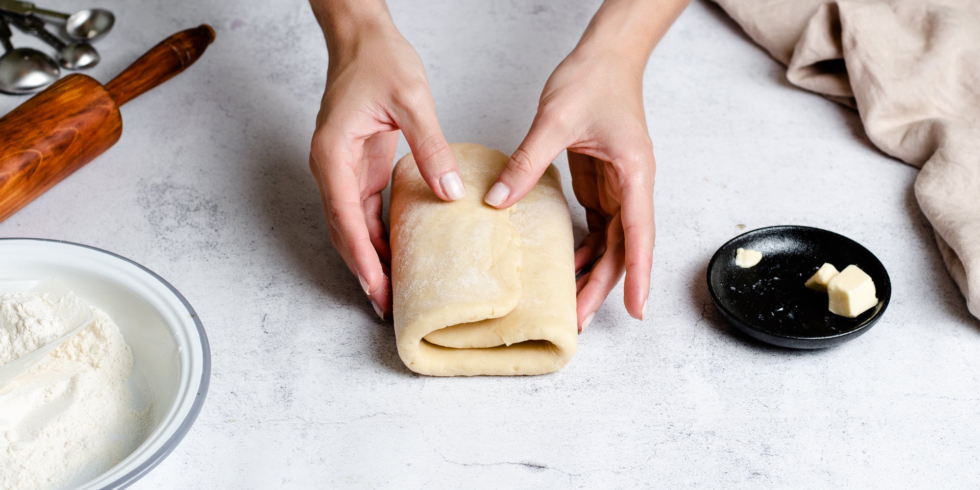 Hands folding Puff Pastry with layers of butter, ready to roll out. Next to a dish of butter and a bowl of flour