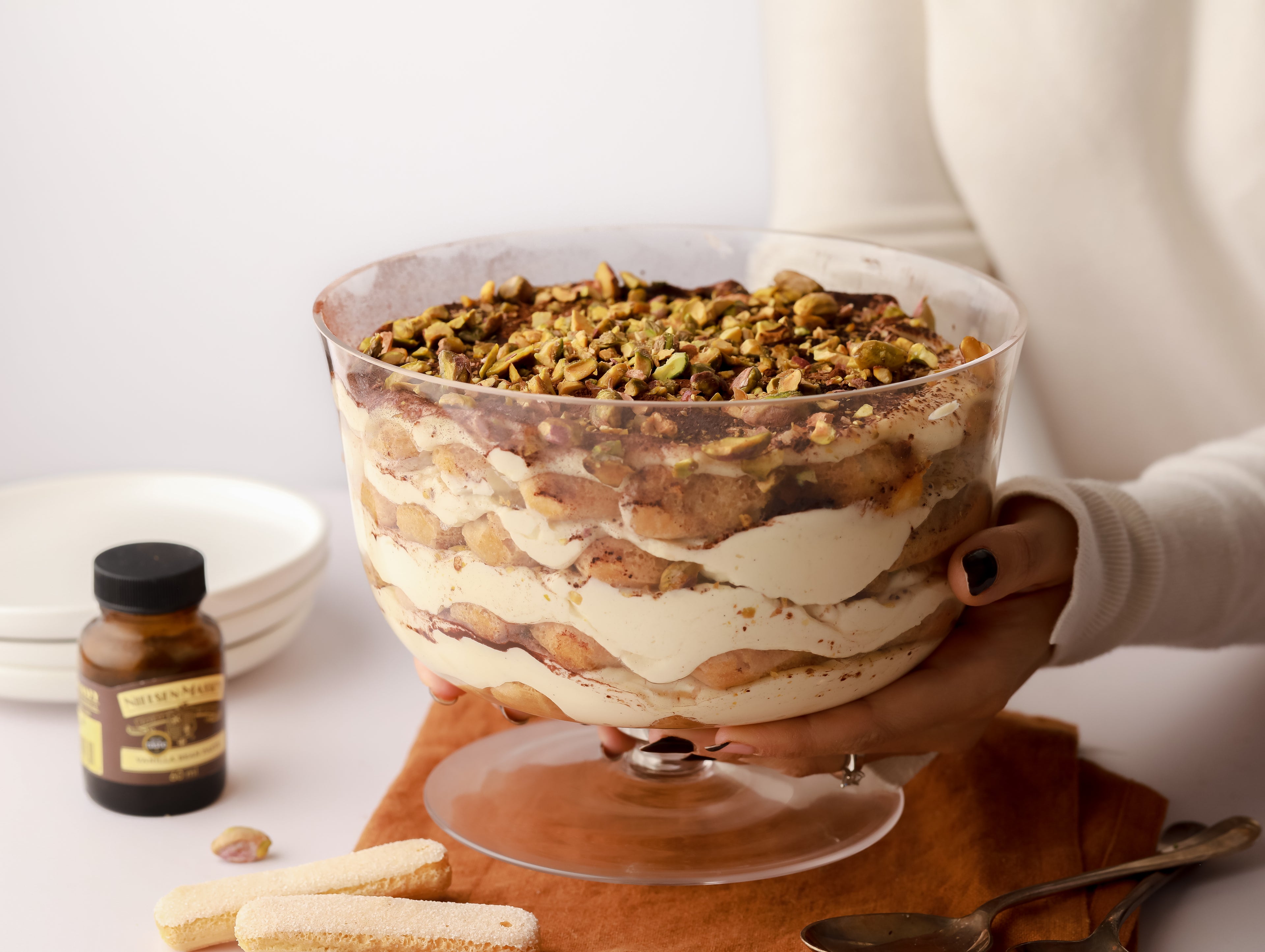 Large bowl of trifle being held by two hands beside a jar of vanilla bean paste