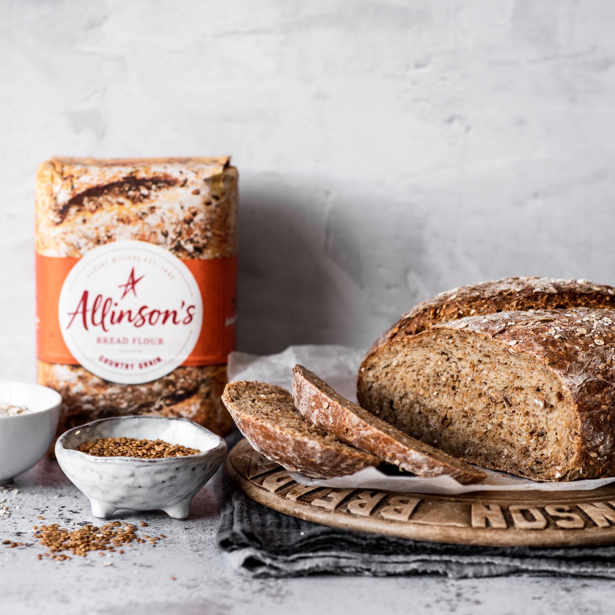 Loaf of bread on bread board with two slices cut infront. Bowl of linseed and bread flour