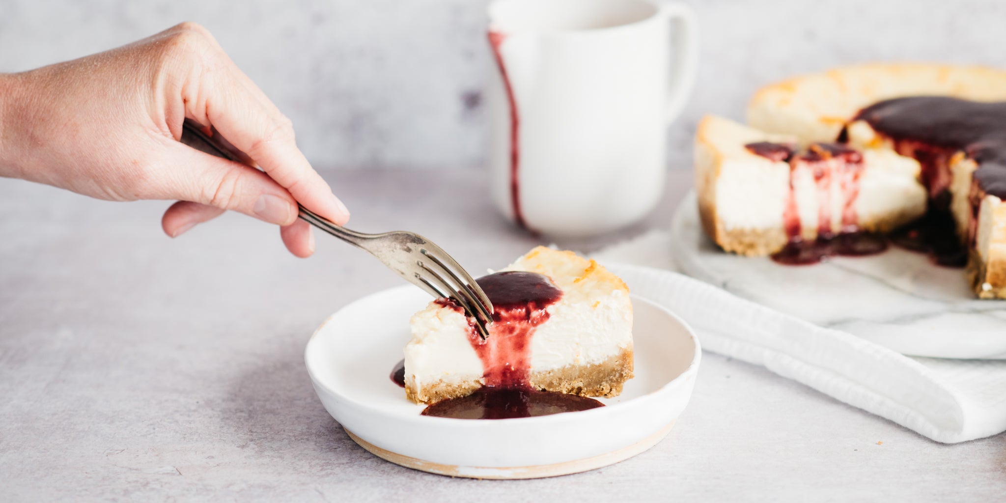 A hand holding a fork slicing into a piece of baked cheesecake covered in raspberry sauce