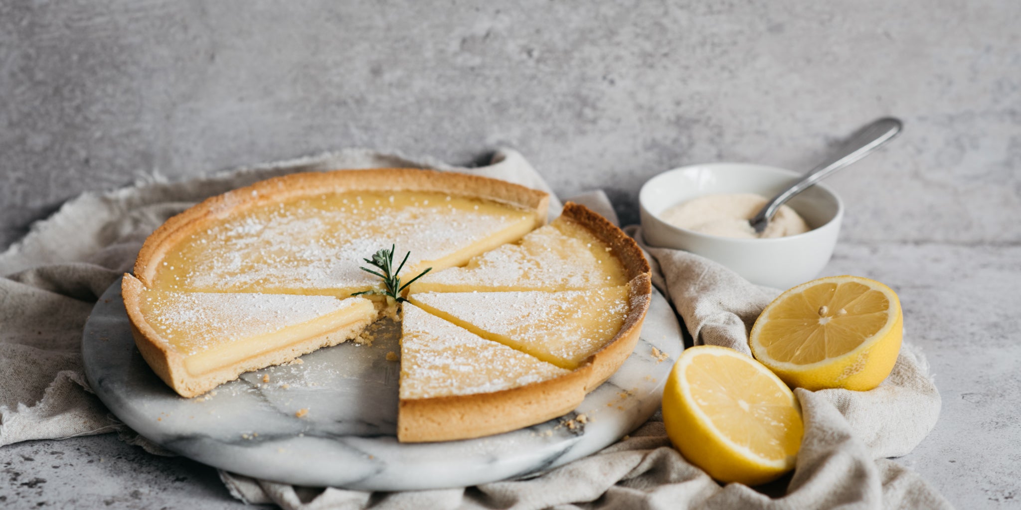 Lemon Tart sliced into servings, on a marble cake board next to slice lemon and a bowl of sugar