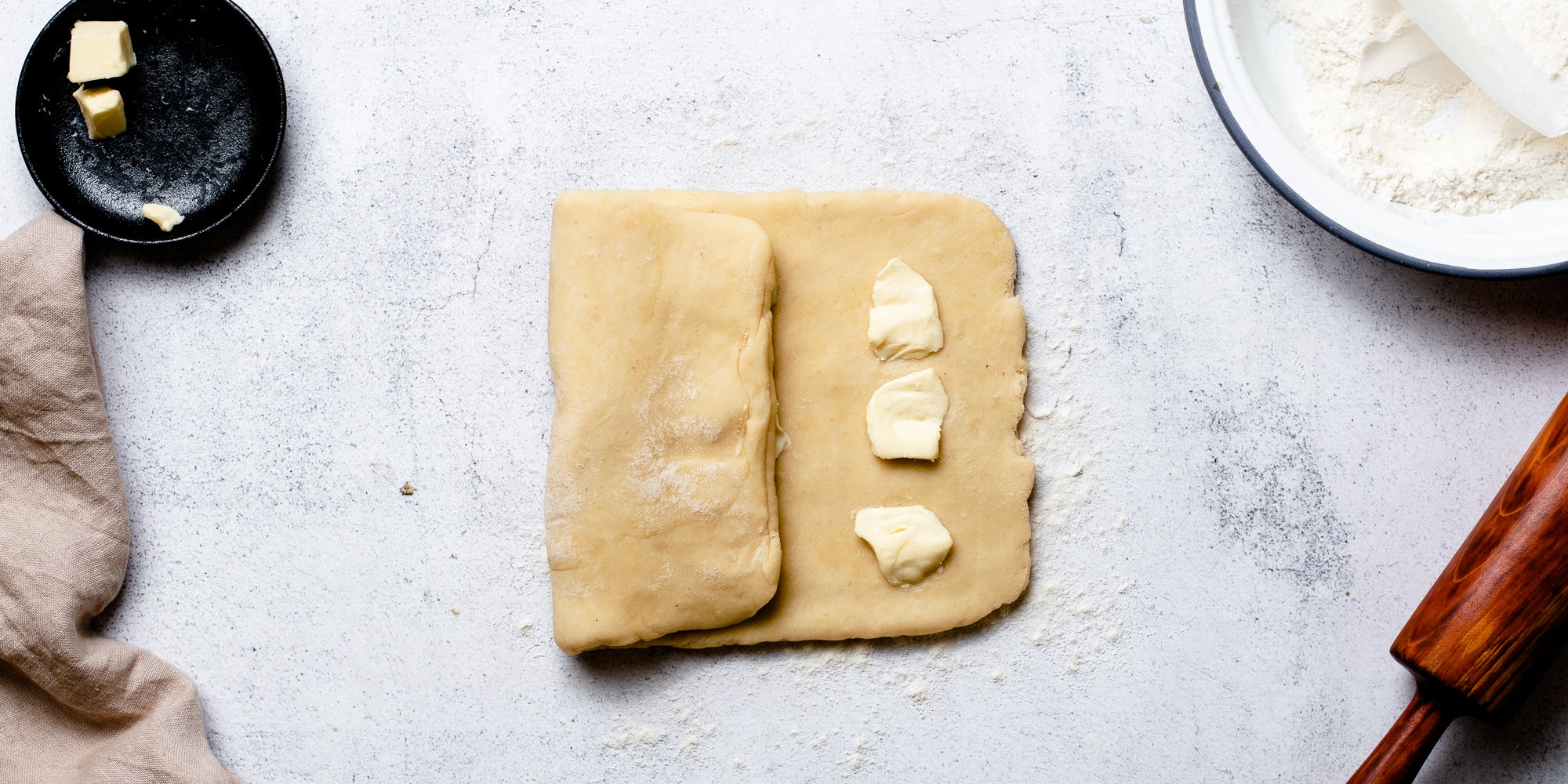 Puff Pastry rolled out with knobs of butter ready to fold into the dough. Next to a rolling pin, bowl of flour and dish of butter