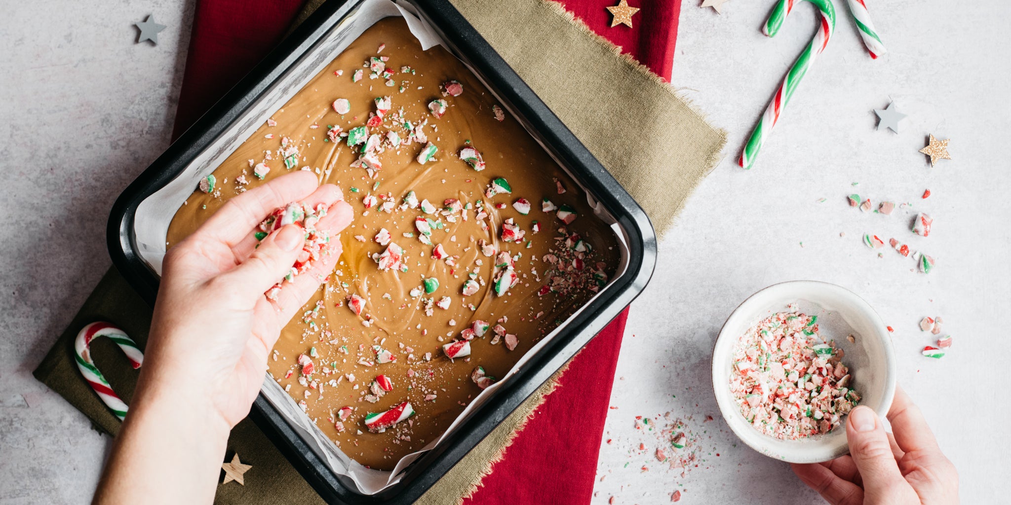 Top view of Candy Cane Fudge being sprinkled with chopped candy cane by a hand. In a tray lined with parchment paper on a red linen cloth