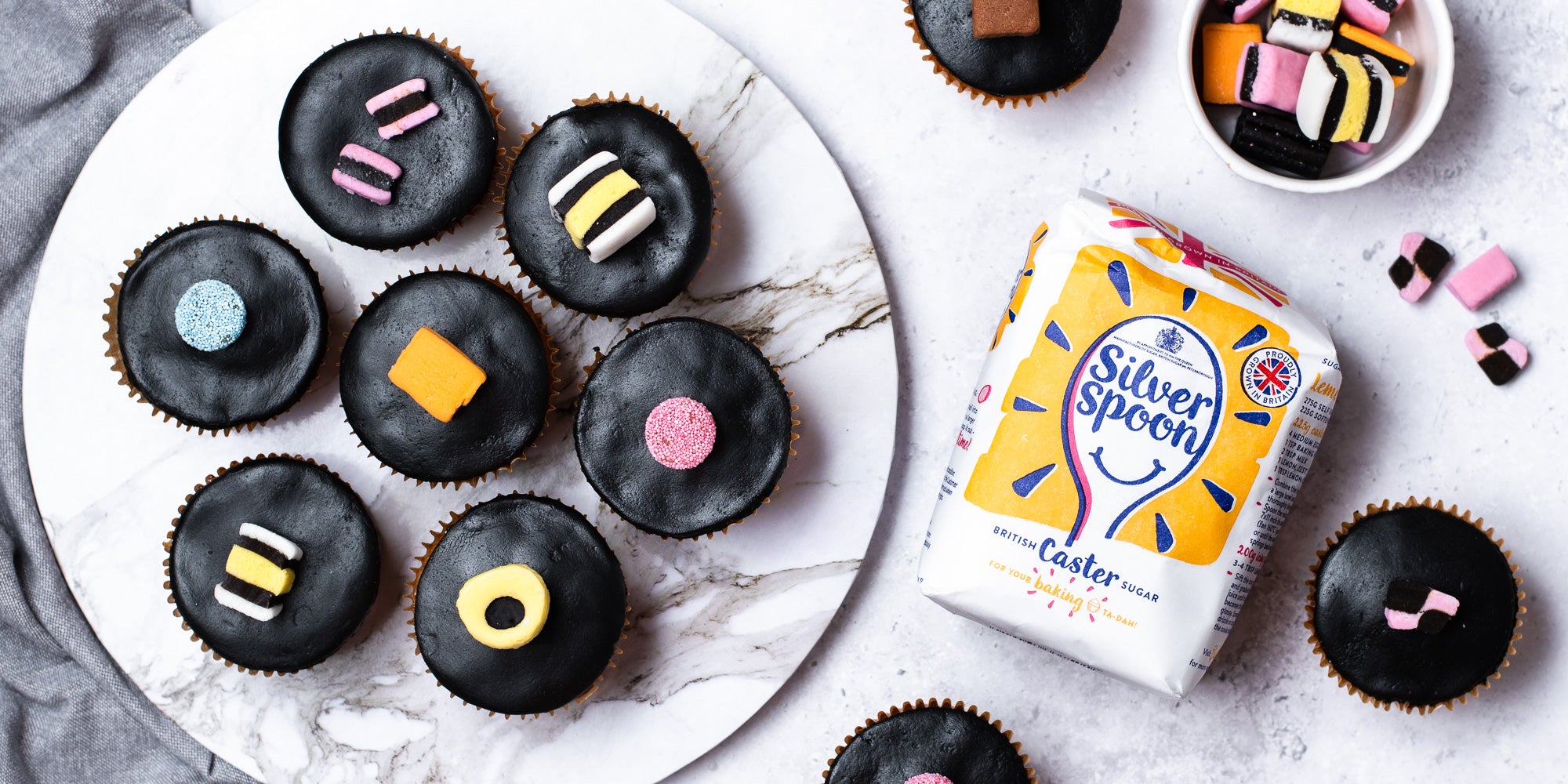 Overhead shot of cupcakes in brown cases, black fondant, topped with liquorice allsorts and sugar pack