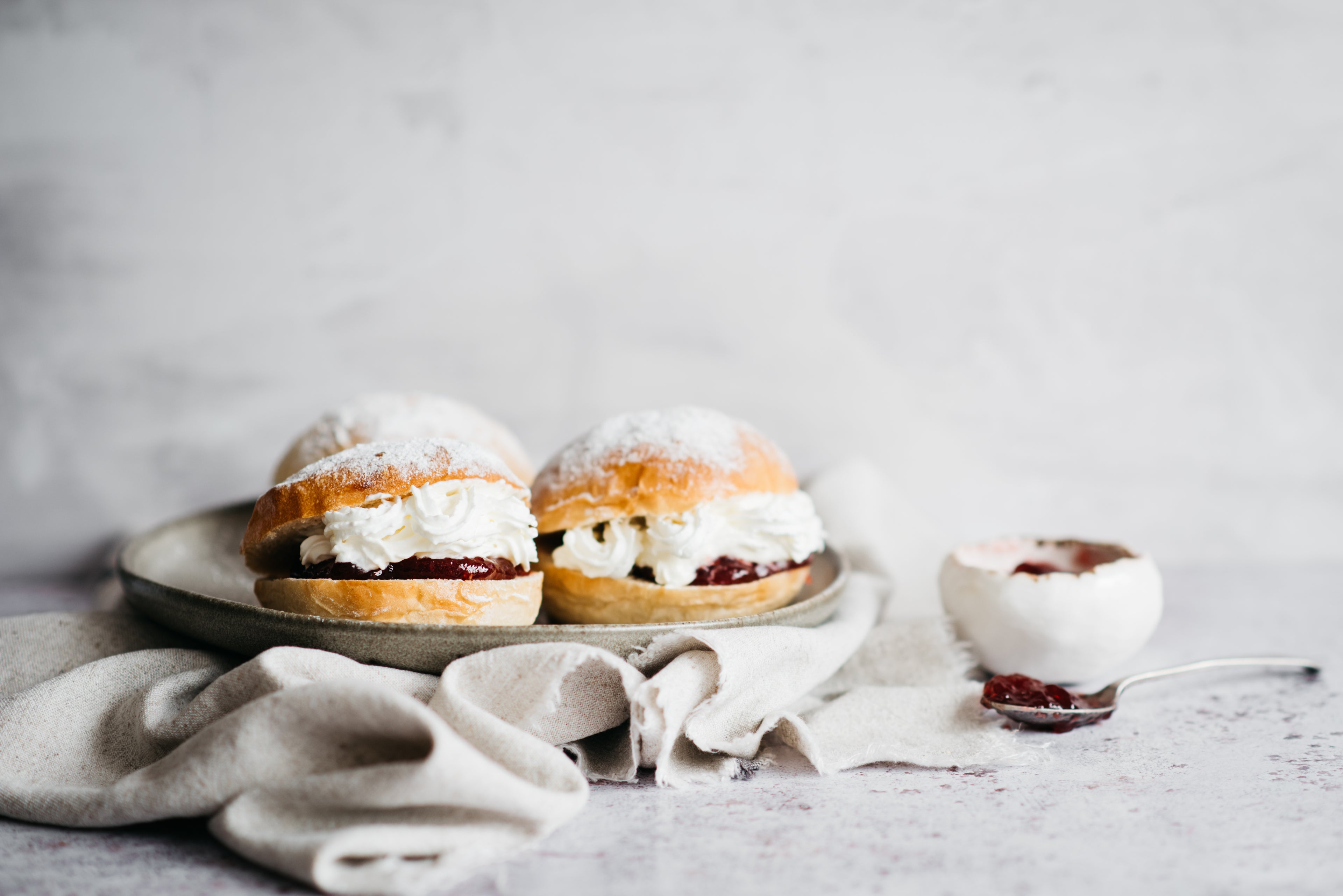 Close up of two Devonshire Splits on a plate, next to a bowl of jam with a spoonful of jam next to it