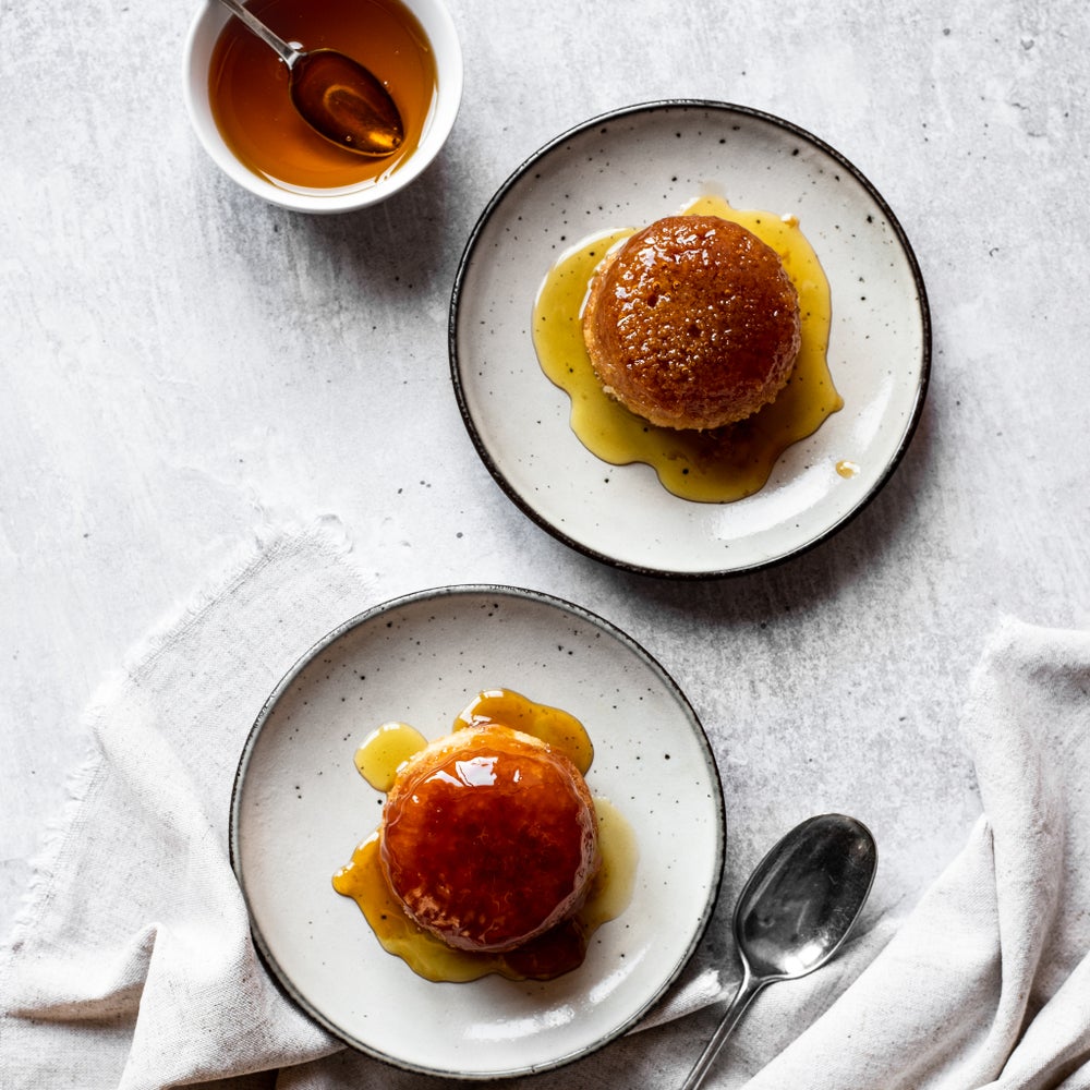 Two saucers each with a mini treacle sponge cake dripping with golden syrup on a grey background, shot from above.