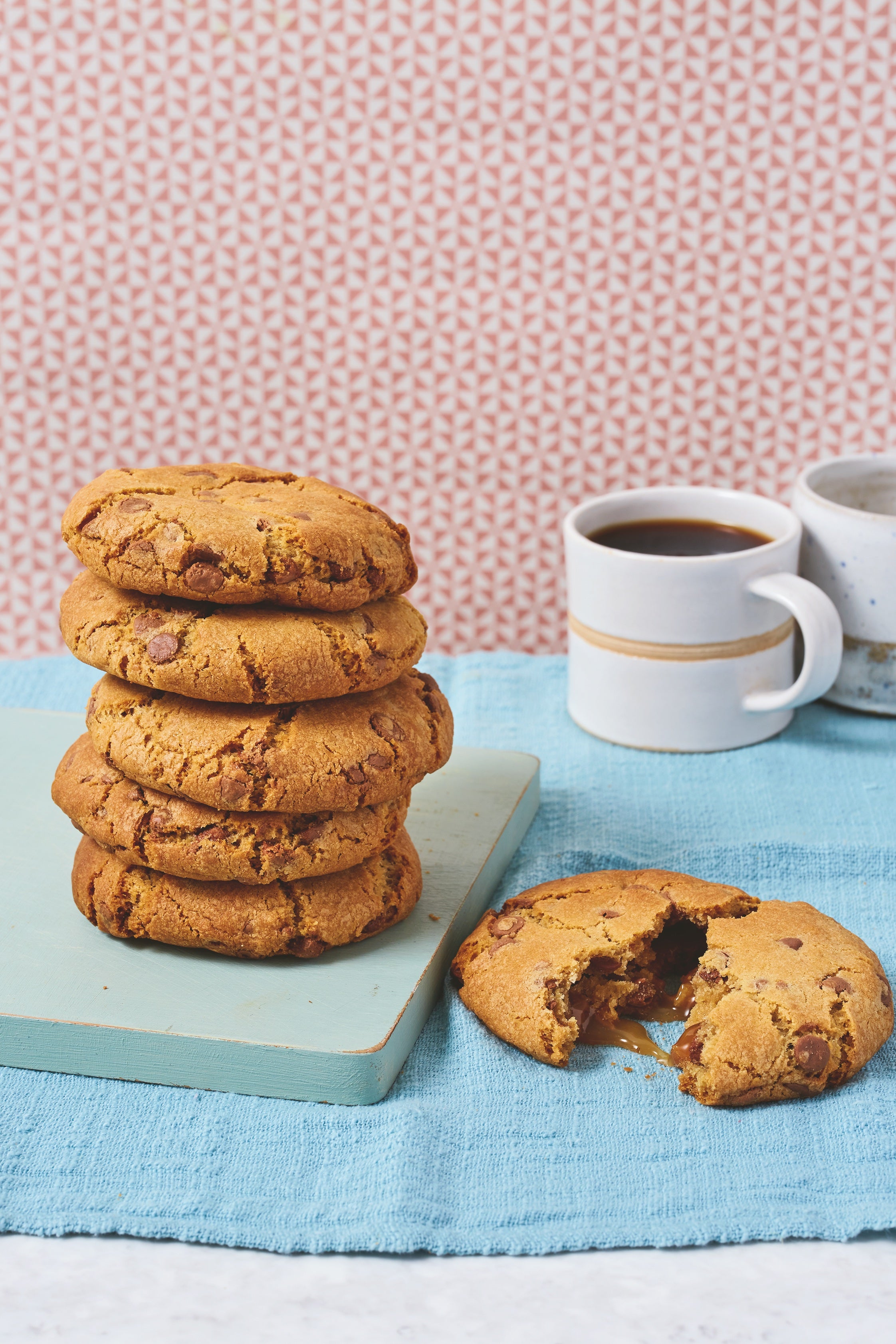 Stack of 5 cookies and one pulled open in front with caramel centre. Cup of coffee in background