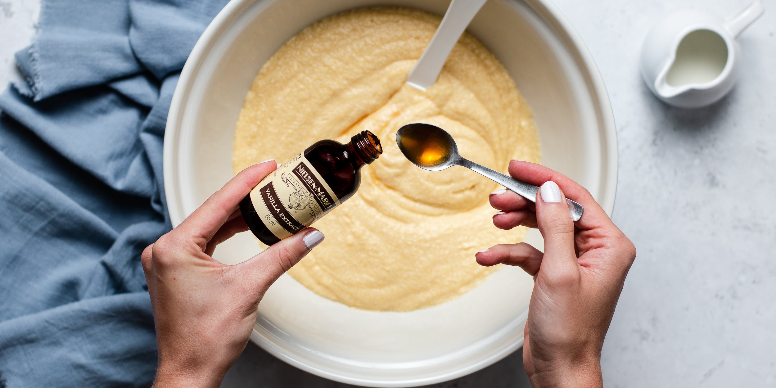 Hand pouring vanilla extract into a spoon over a bowl of cake mixture