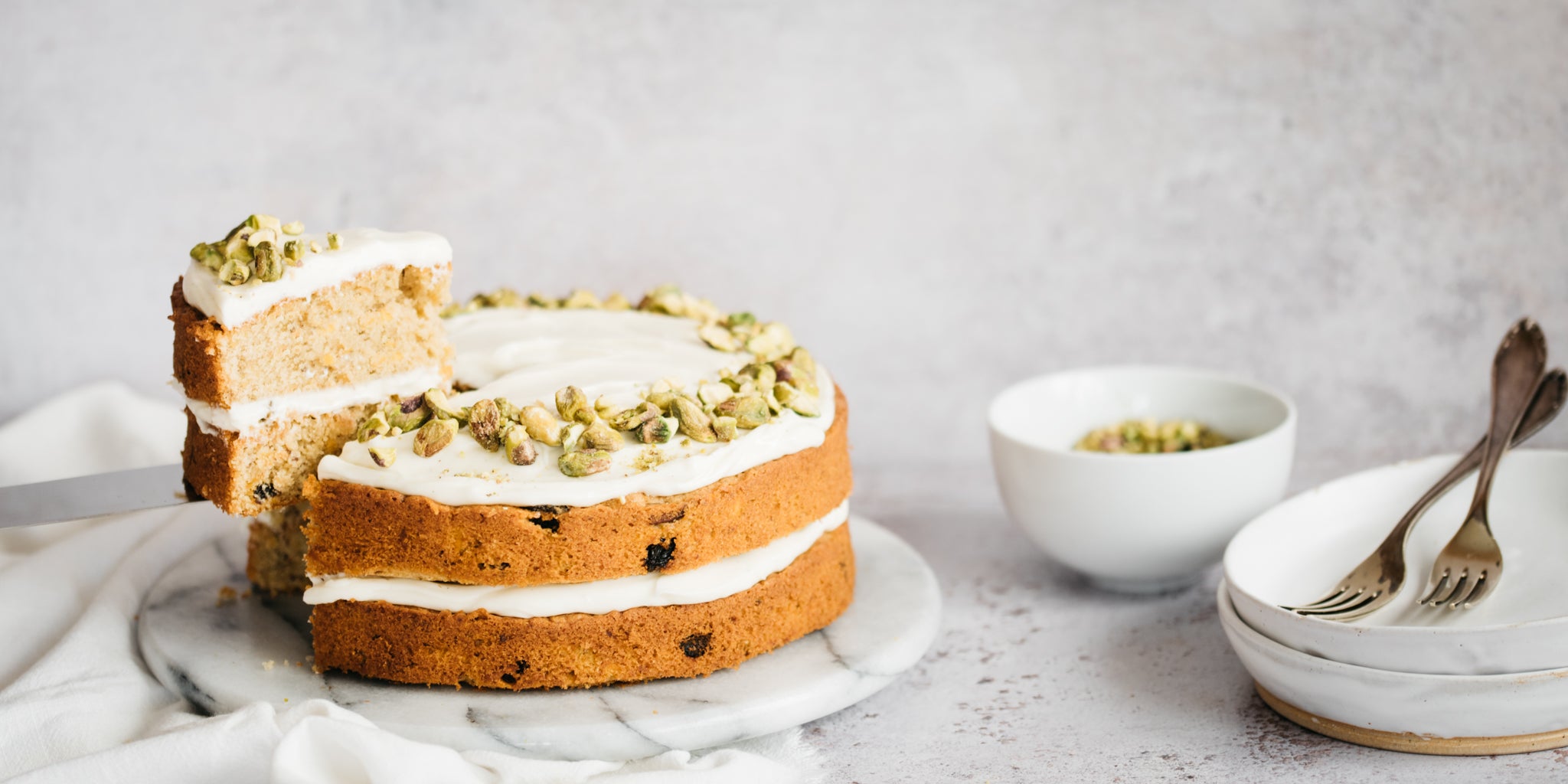 Carrot cake on marble plate with a slice removed. Bowl of nuts next to a stack of white plates and forks