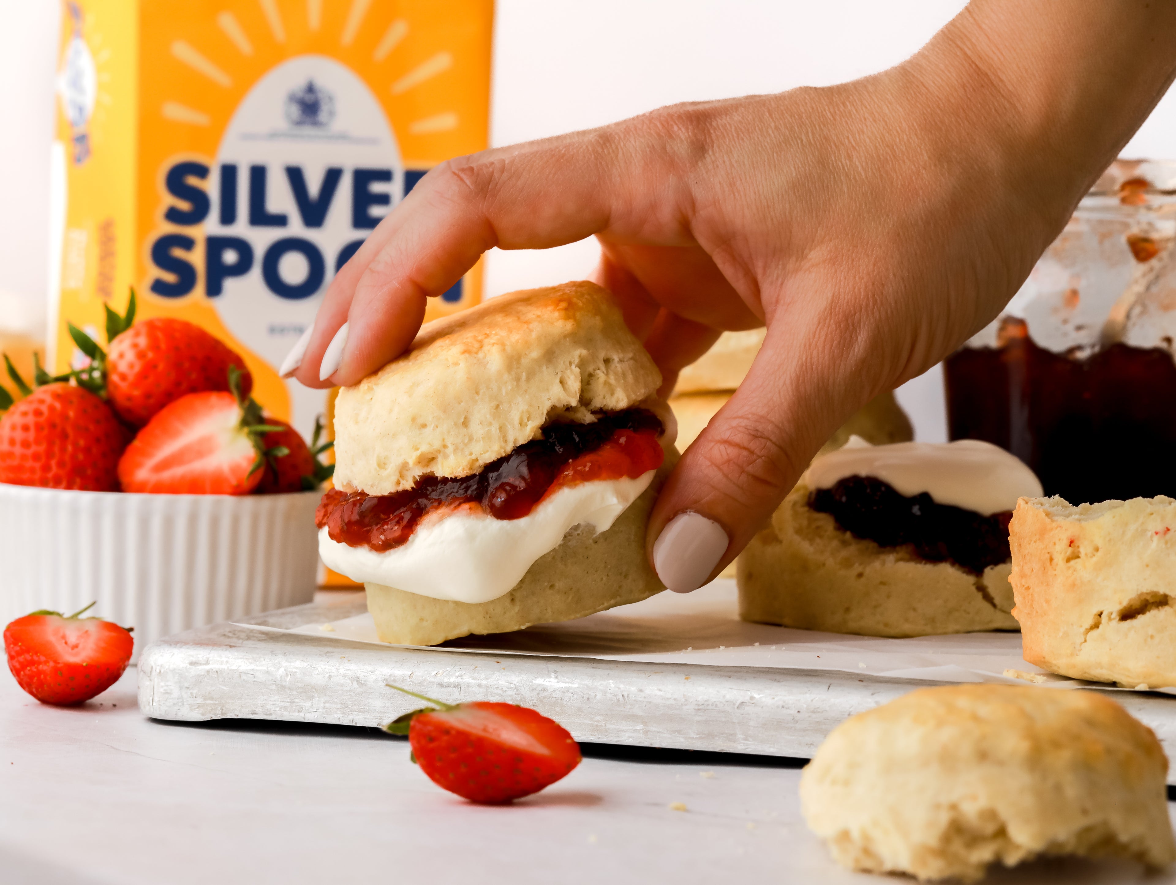 Hand holding homemade scone filled with with jam and cream next to a bowl of strawberries and sugar