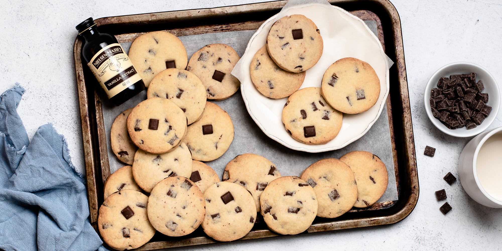 A batch of Chocolate Chunk Shortbread on a baking tray, next to a bottle of Nielsen-Massey extract and a bowl of chocolate chunks