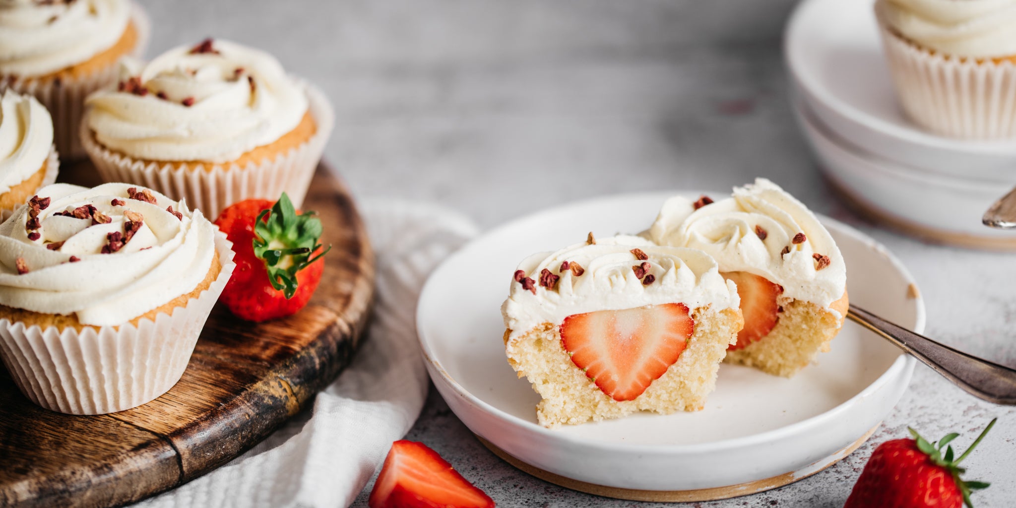 Strawberry cupcake cut in half showing a strawberry centre and topped with buttercream. More cupcakes on the plate to the side