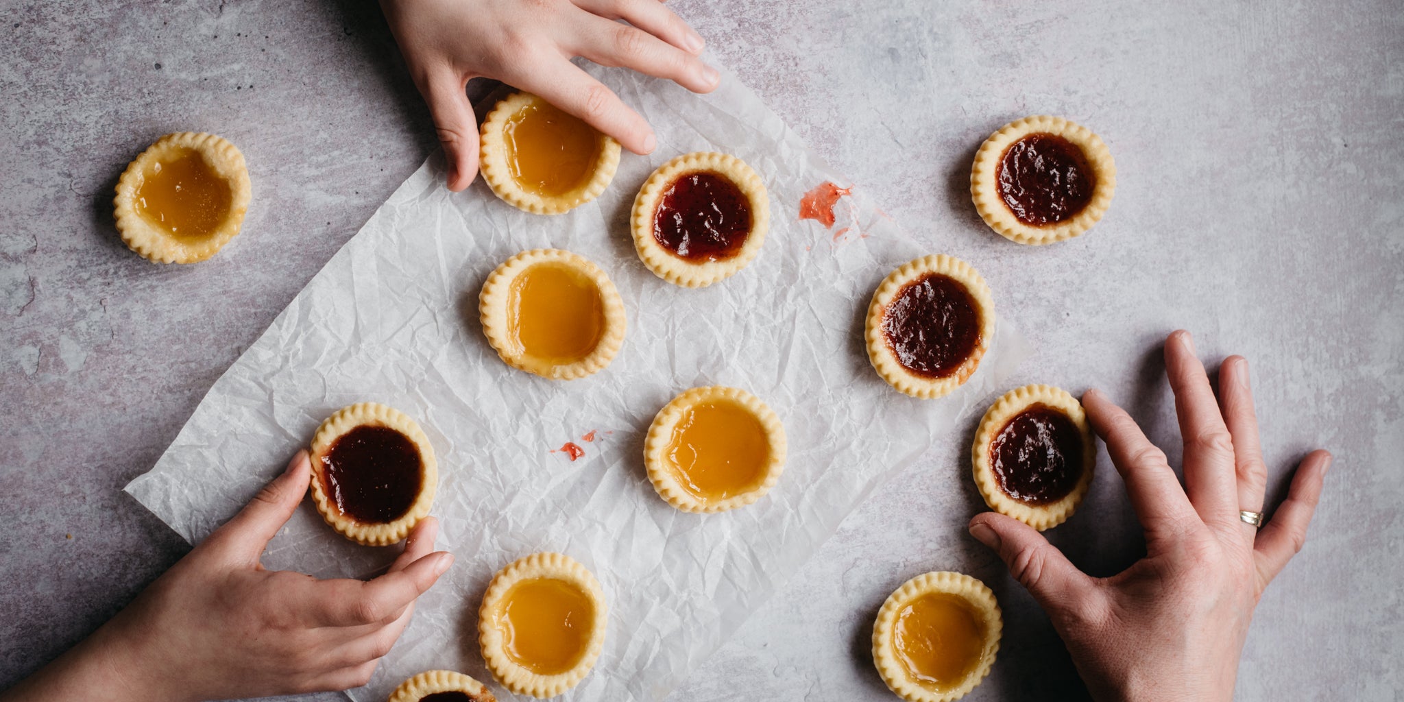 Hands reaching for a jam tart, with Apricot & Strawberry Jam Tarts served on parchment paper