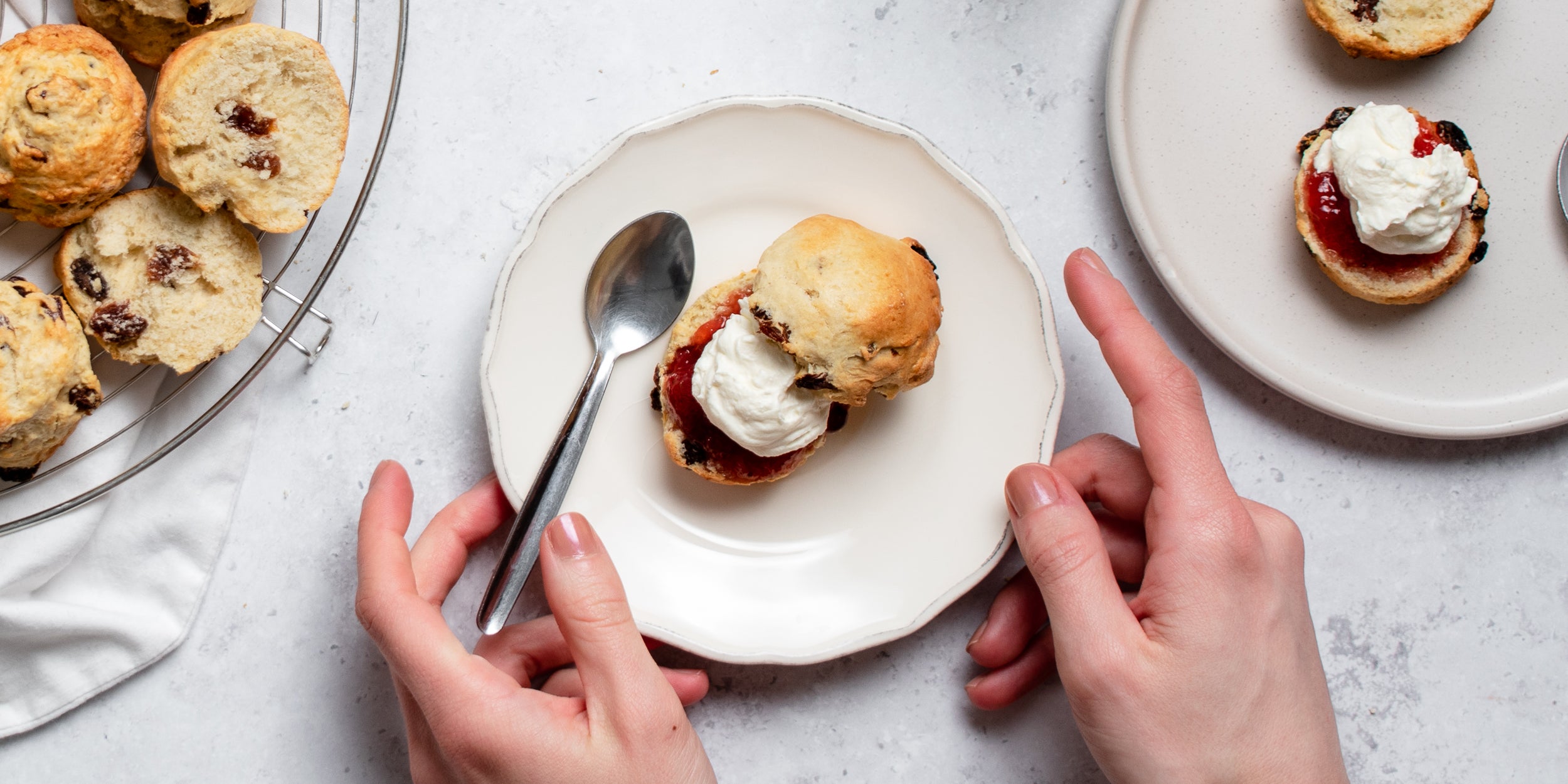 Top view of Fruit Scone sliced in half with cream and jam, with a hand holding a spoon ready to dig in
