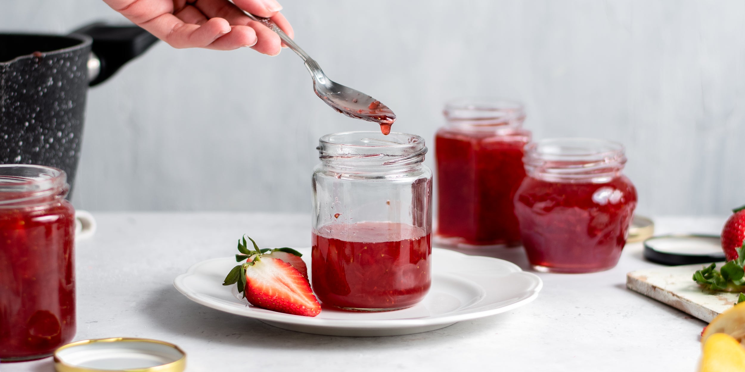 Strawberry Jam being spooned out of a jar with a hand holding a spoon next to sliced strawberries