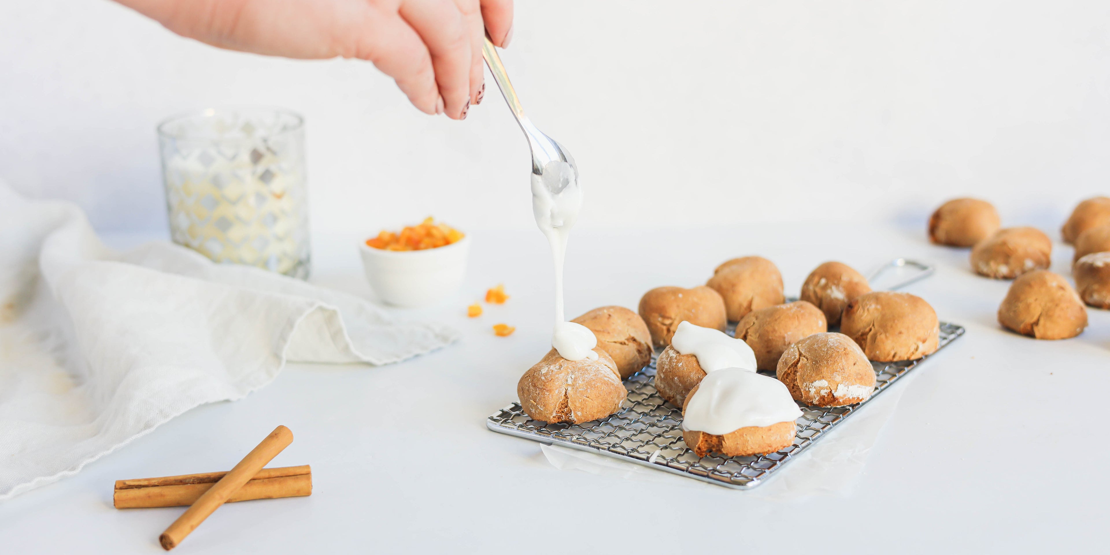 Pfeffernusse being drizzled by a hand with Silver Spoon Icing, on a wire rack, with sticks of cinnamon in the foreground