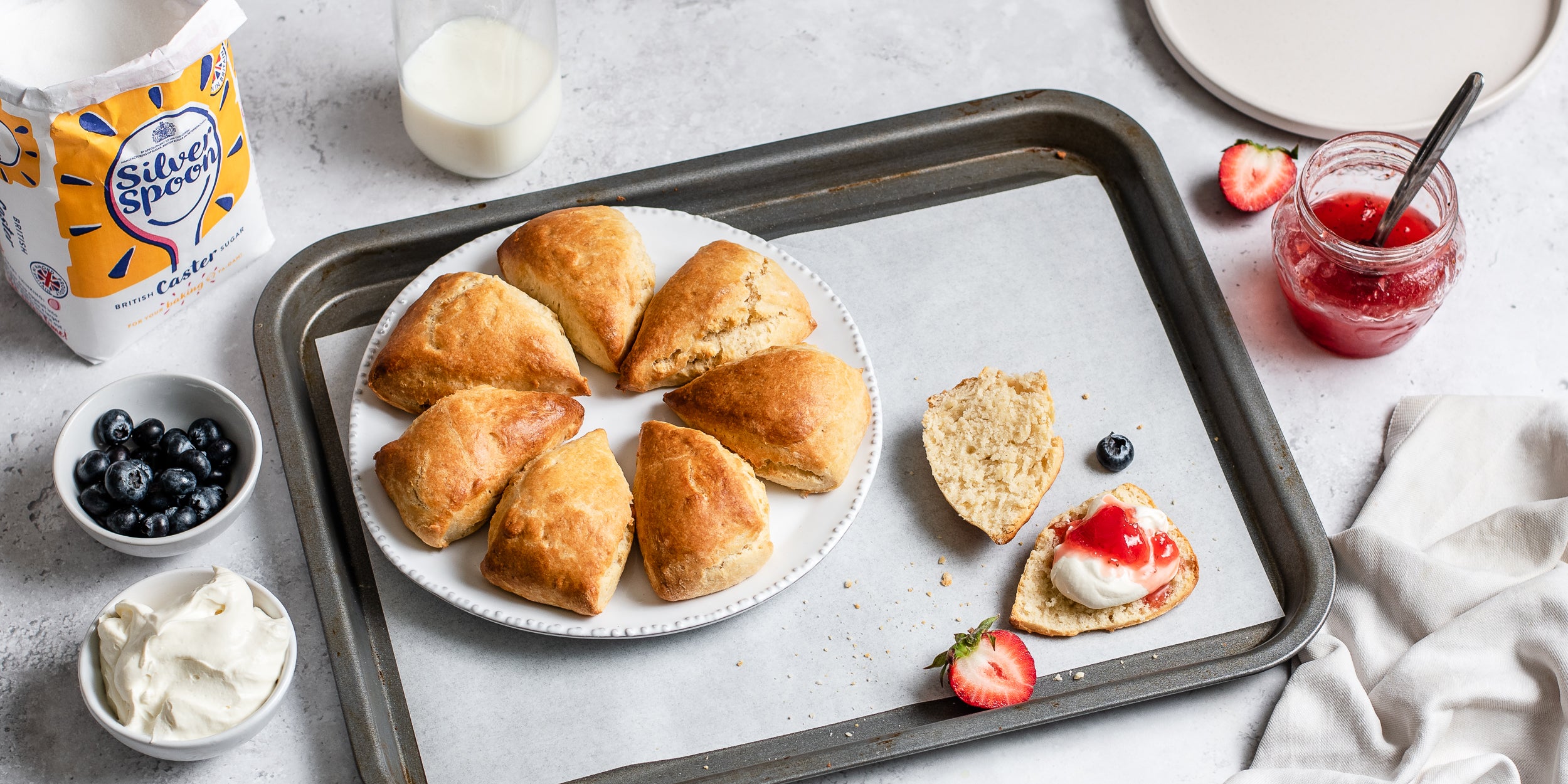 Easy Scones served on a plate, cut into portion and dolloped with cream and jam. Next to a bag of Silver Spoon caster sugar