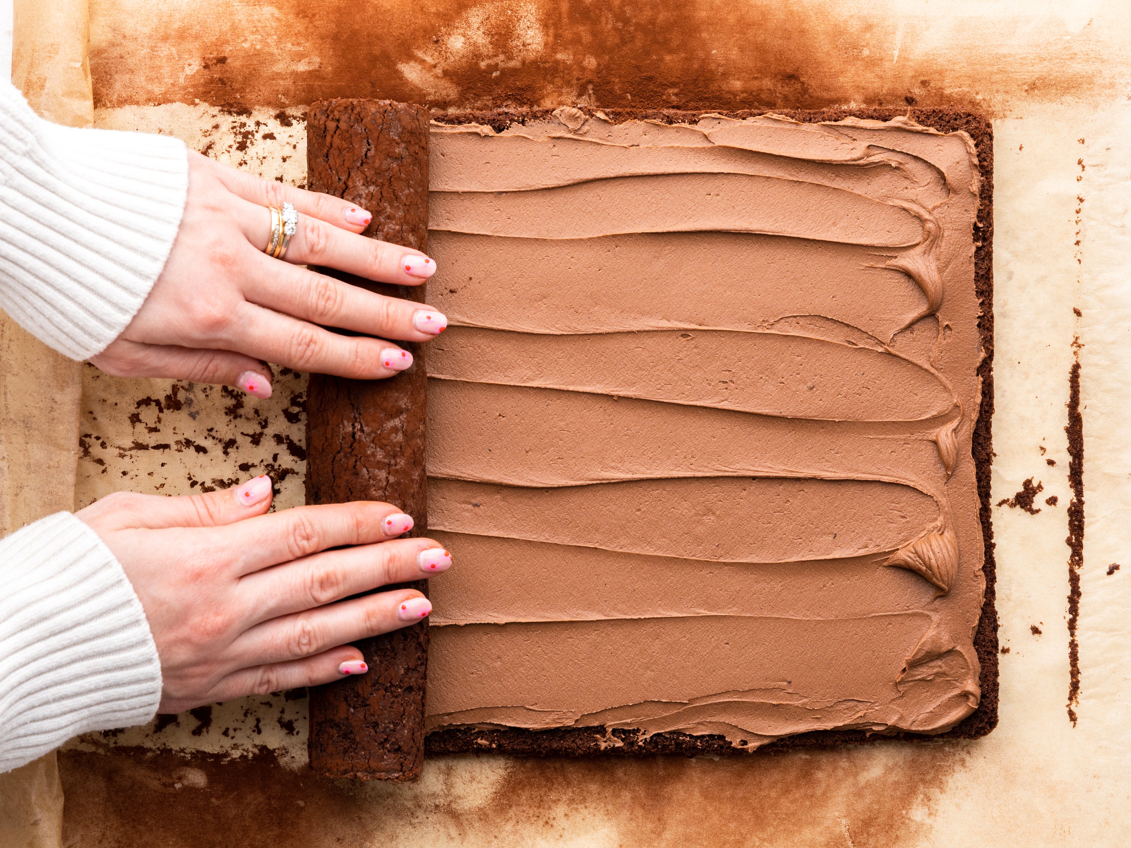 Hands rolling up a swiss roll covered in chocolate buttercream