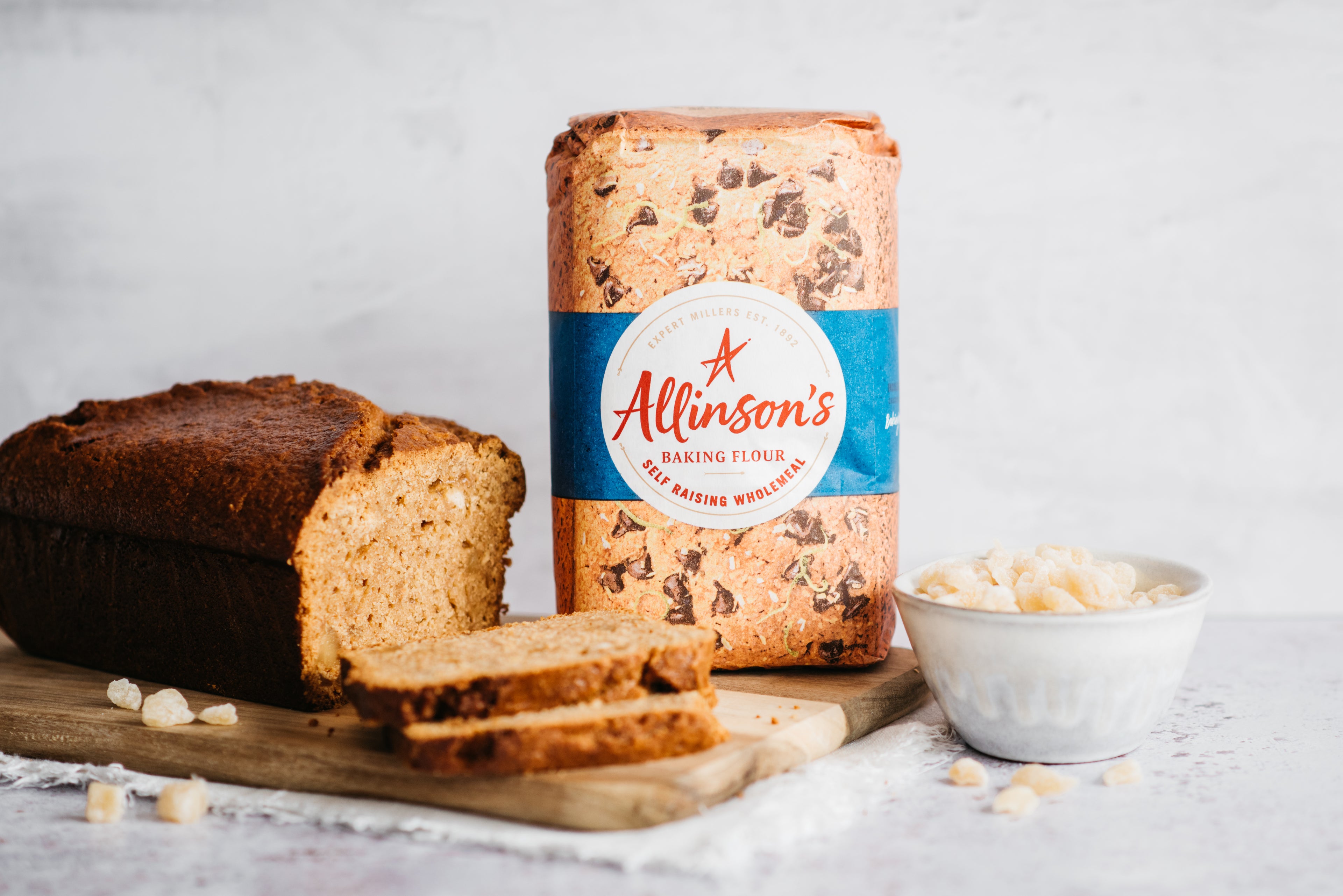 Gingerbread Loaf sliced on a wooden serving board, next to a bag of Allinson's self raising flour and a bowl of crystallised ginger