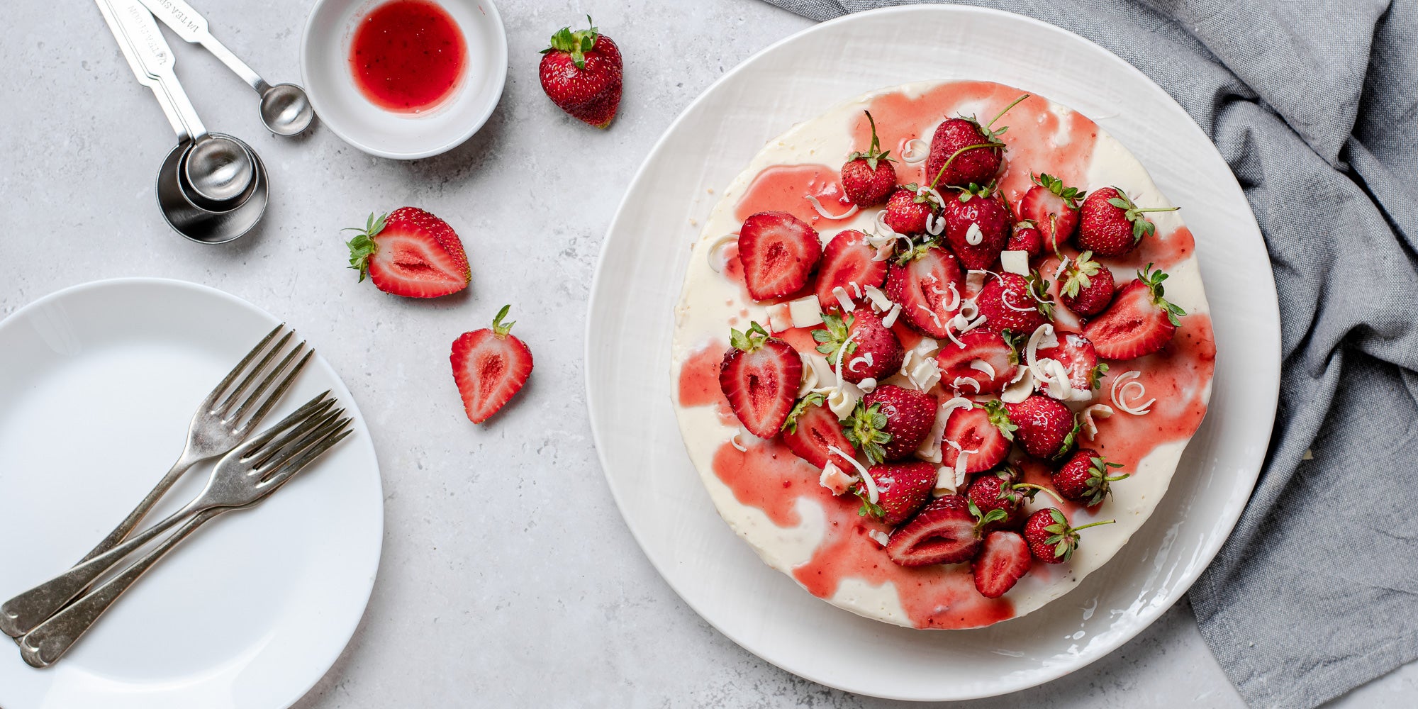 White Chocolate & Strawberry Cheesecake on a plate, next to a stack of plates with forks ready to serve