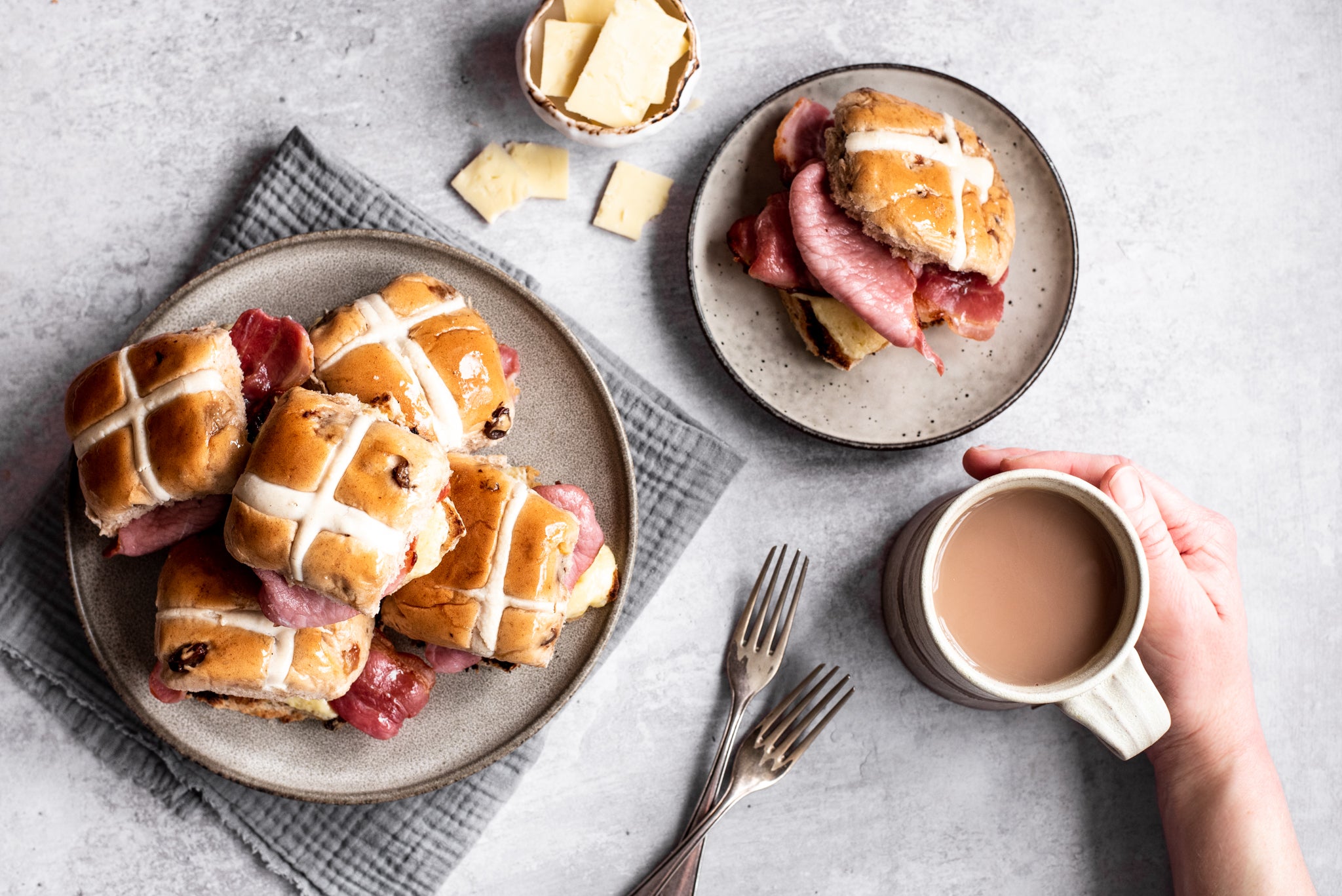 Plate of hot cross buns stacked on top of each other. Plate with hot cross bun and bacon. Cup of tea and hand. Two forks