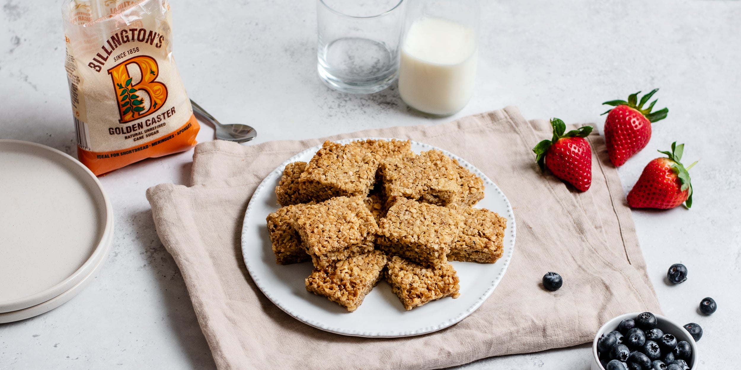 Batch of Easy Flapjacks on a plate, next to fresh berries and a bag of Billington's Golden Caster Sugar