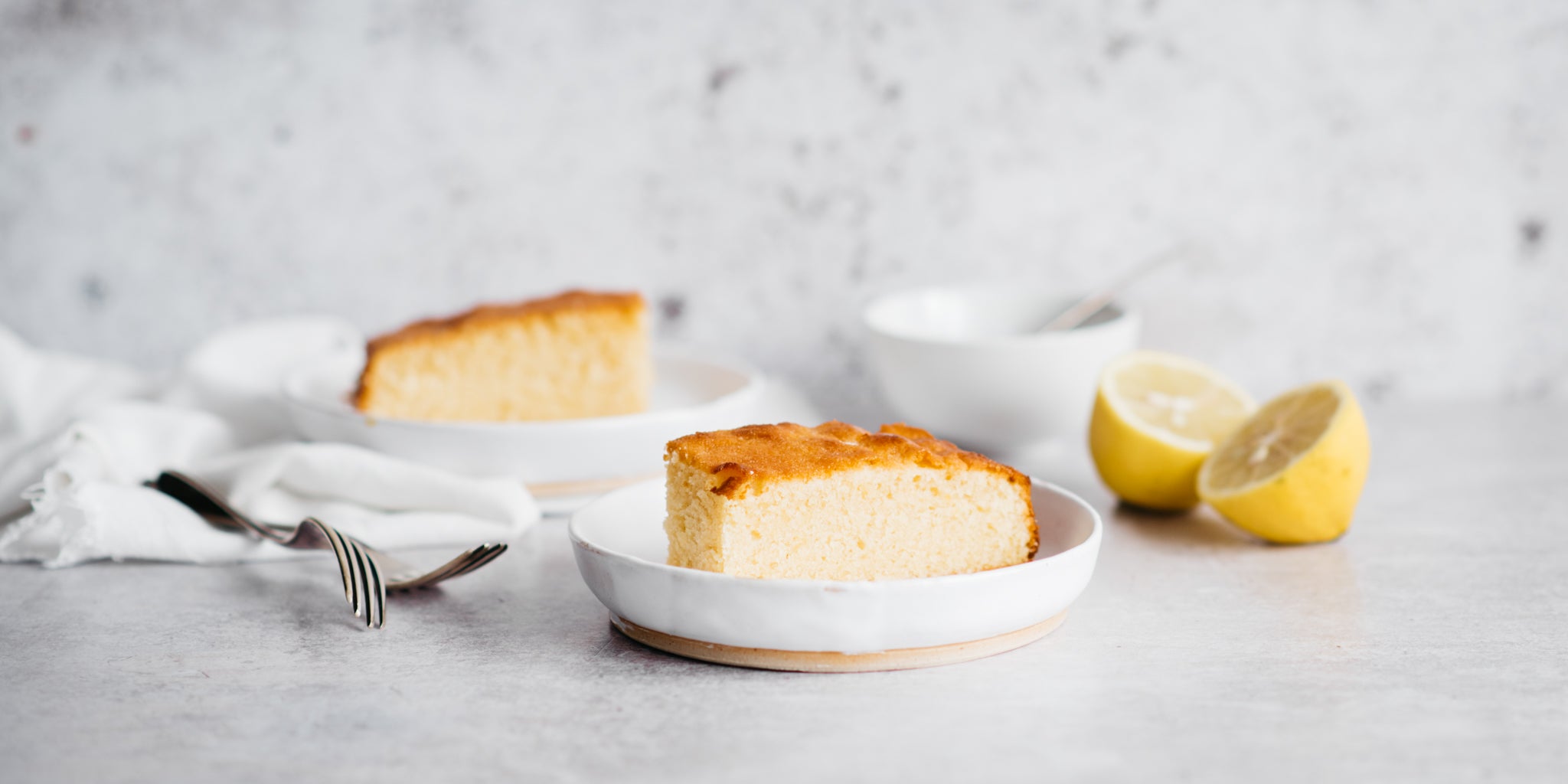 Slice of lemon drizzle on a white plate with sliced lemon beside it