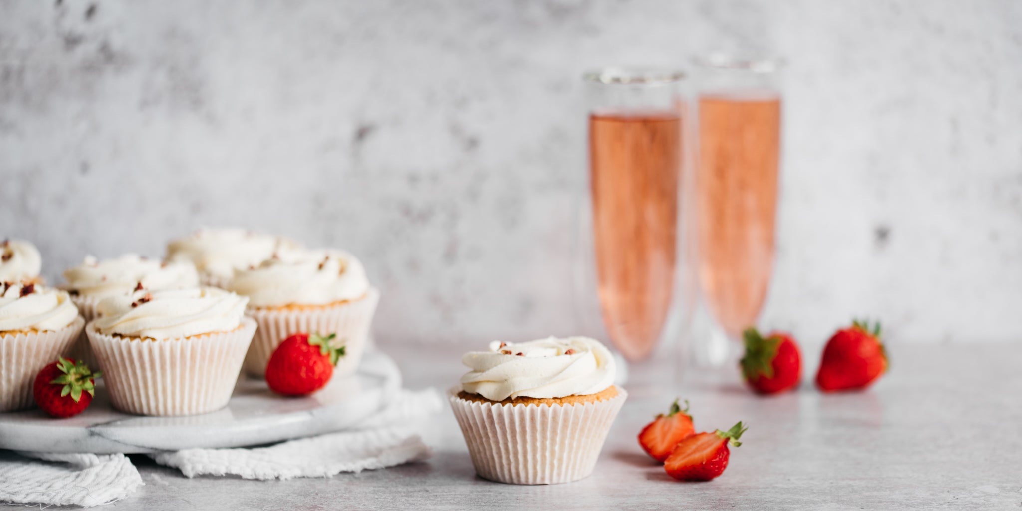 6 cupcakes on a white plate with strawberries. Two glasses of prosecco in the background. One cupcake in the forefront with strawberries around it
