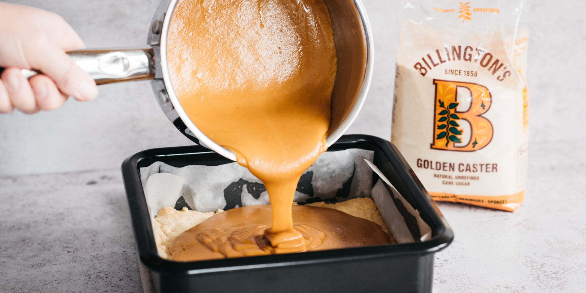 Saucepan of caramel being poured into a baking tin with a pack of sugar beside it