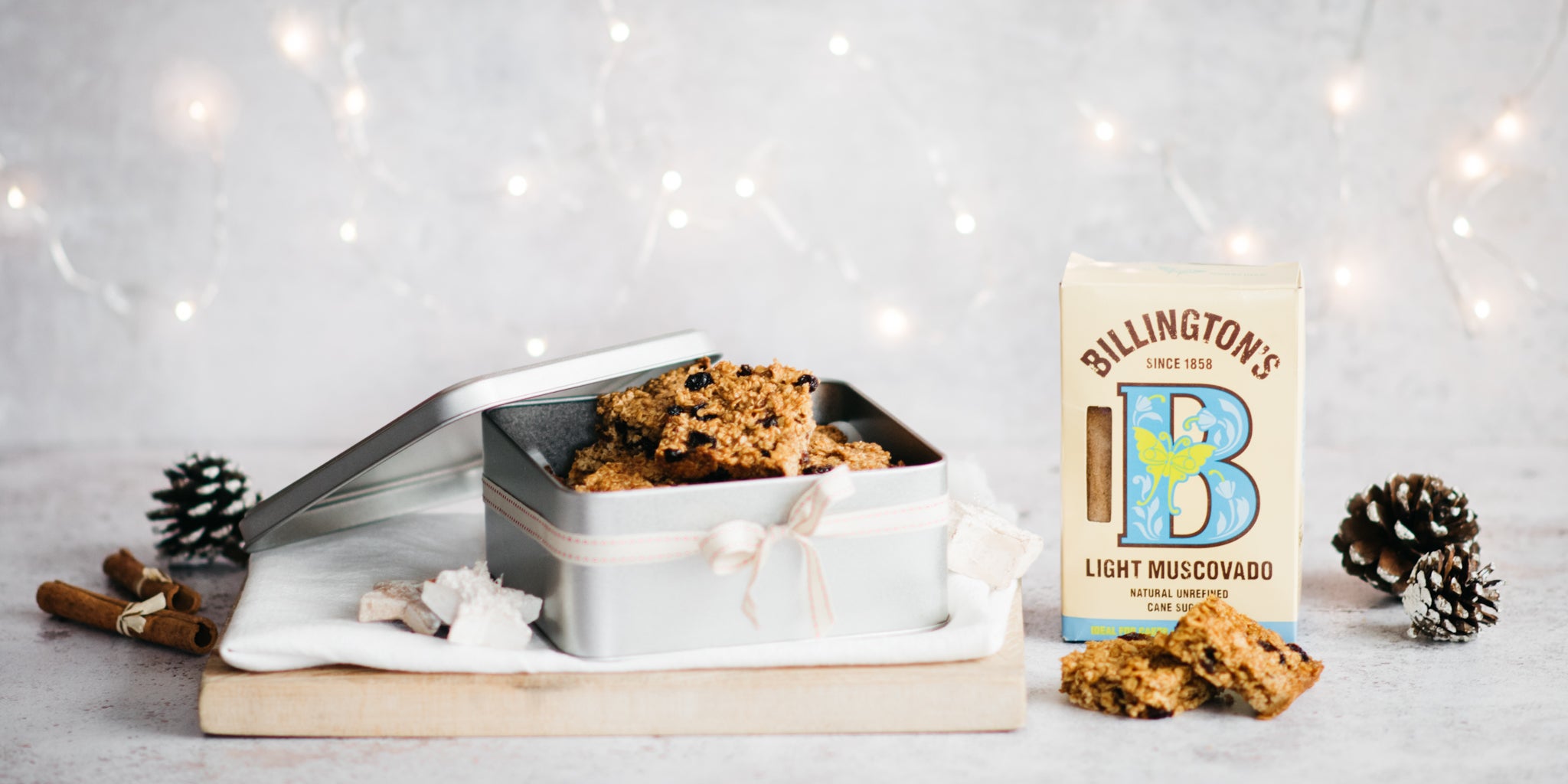 Tin of flapjacks with lid ajar next to a pack of sugar and pine cones. Festive fairy lights in background