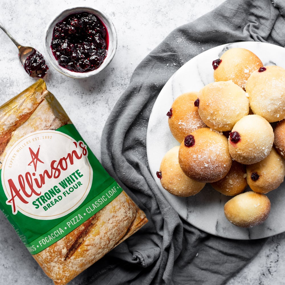 A bag of Allinson's strong white bread flour laid flat next to a marble round plate piled with baked jam doughnuts. A small dish of red jam is next to them both.