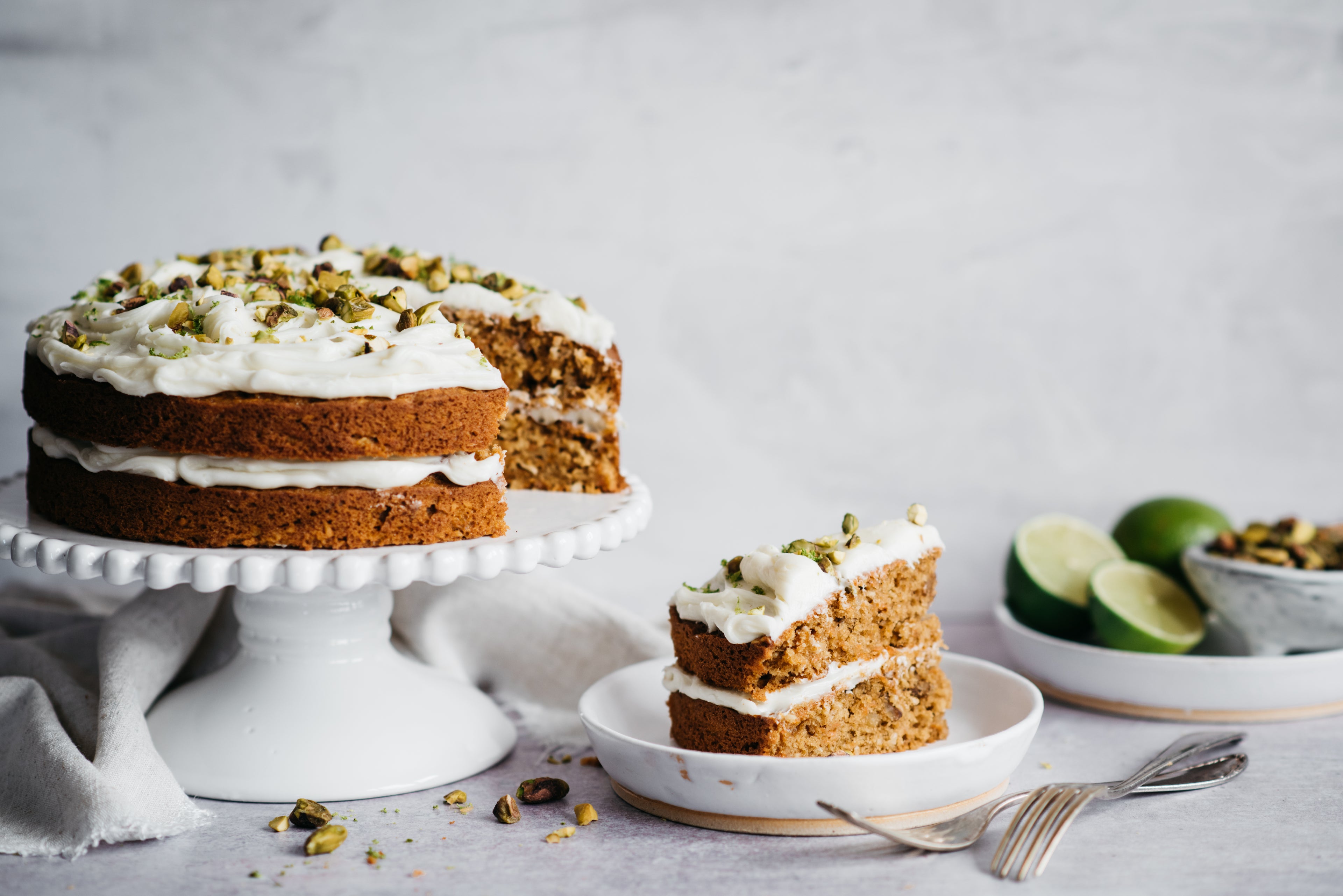 Carrot cake with icing on cake stand and cake slice on plate. Limes in the background