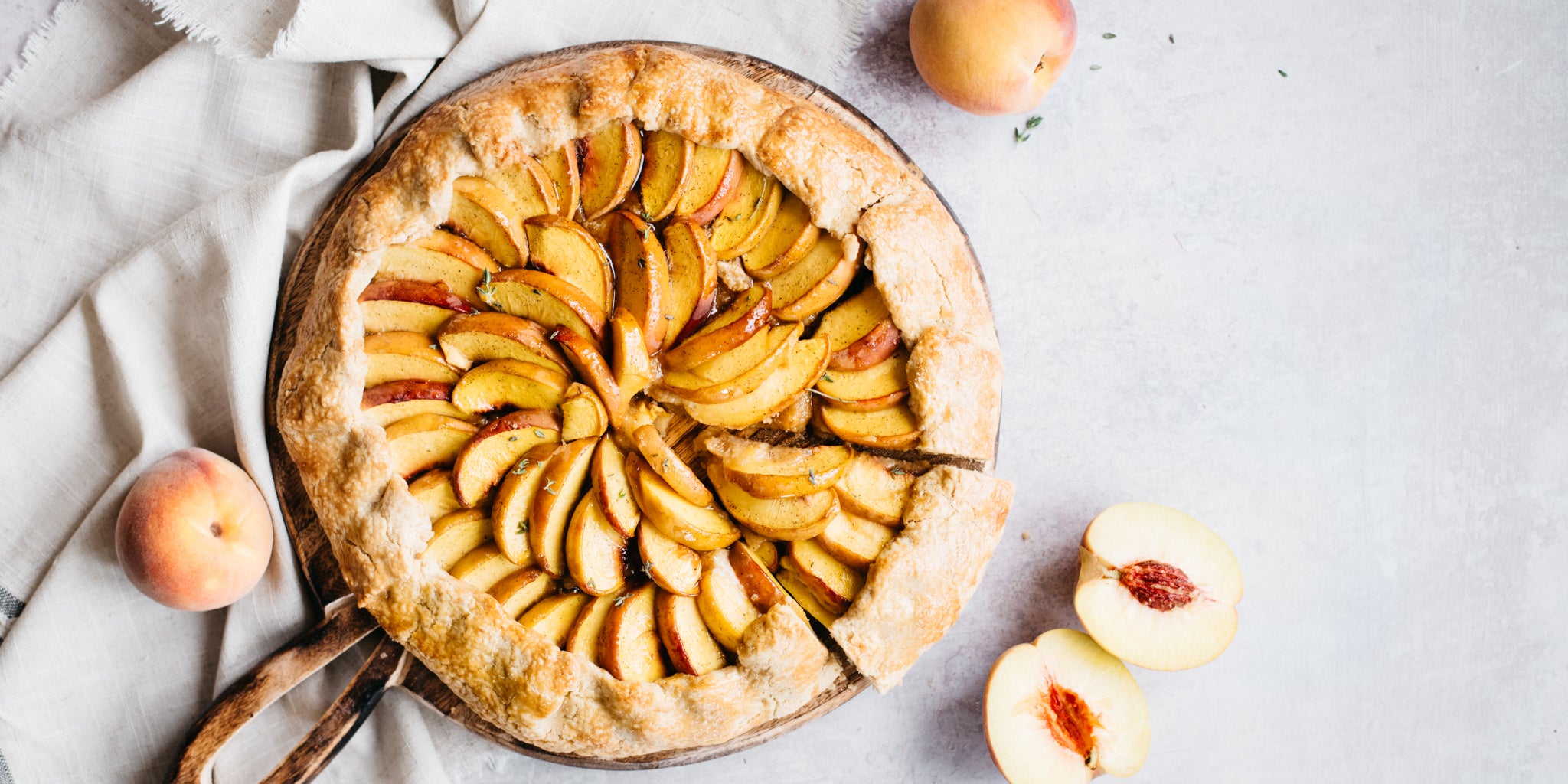Peach Galette on a circular board with a slice cut. Peaches beside, one sliced in half
