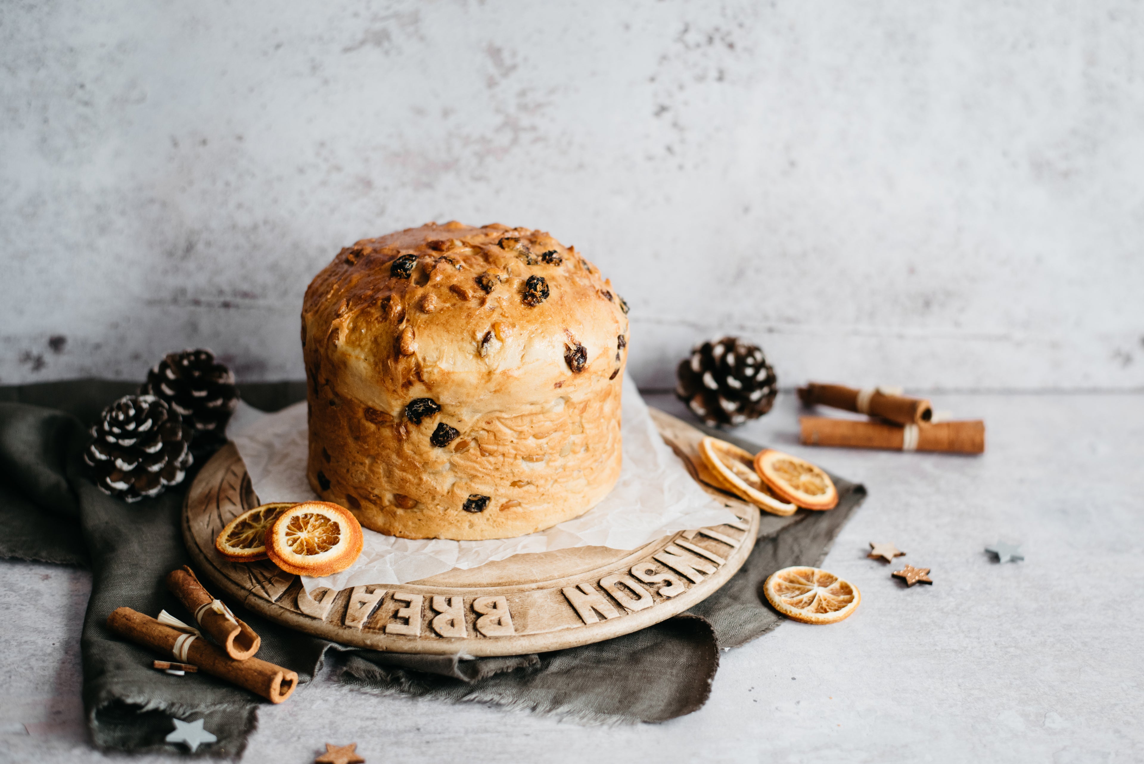 Panettone served on a wooden serving board, next to cinnamon sticks, dried orange and pine cones