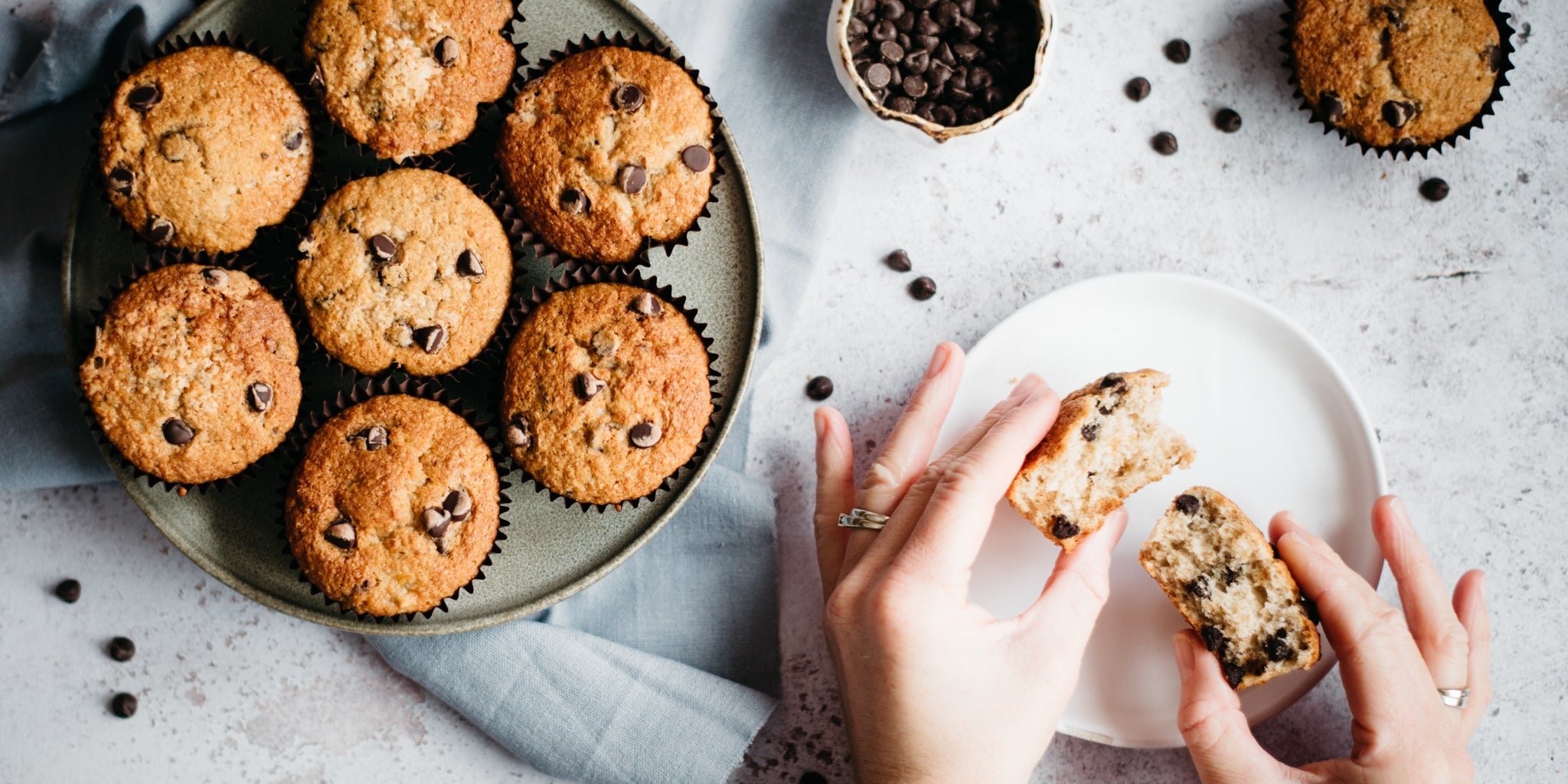 Top view of Wholemeal Banana & Chocolate Chip Muffins on a wire cooling rack, with a hand tearing a muffin apart showing the chocolate chip filled insides