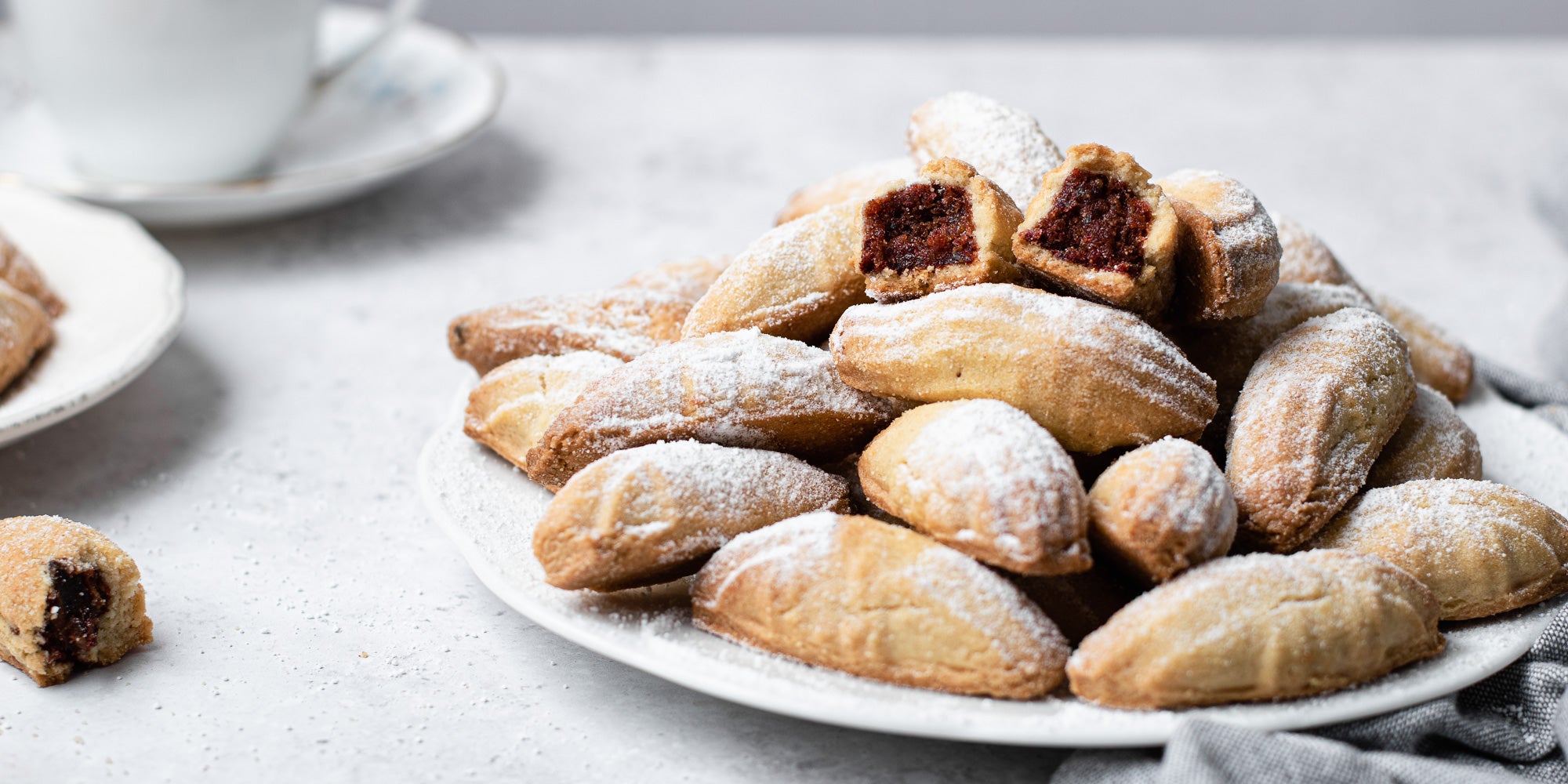 A plate of Mamoul dusted with icing sugar next to a cup of tea