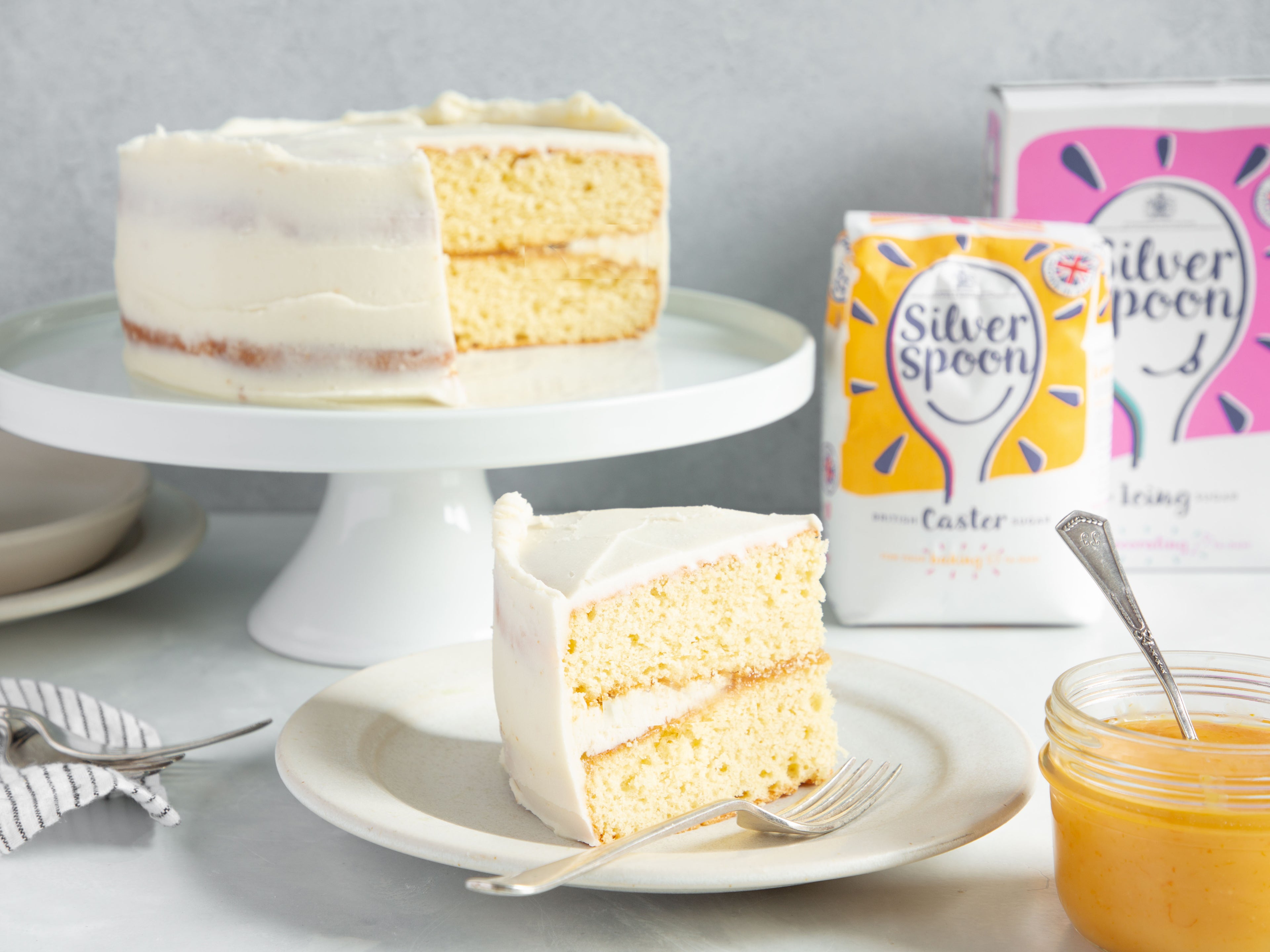 Classic Lemon Cake on a cake stand with a slice cut out showing the lemon filling next to a bag of Silver Spoon Icing and Caster sugar