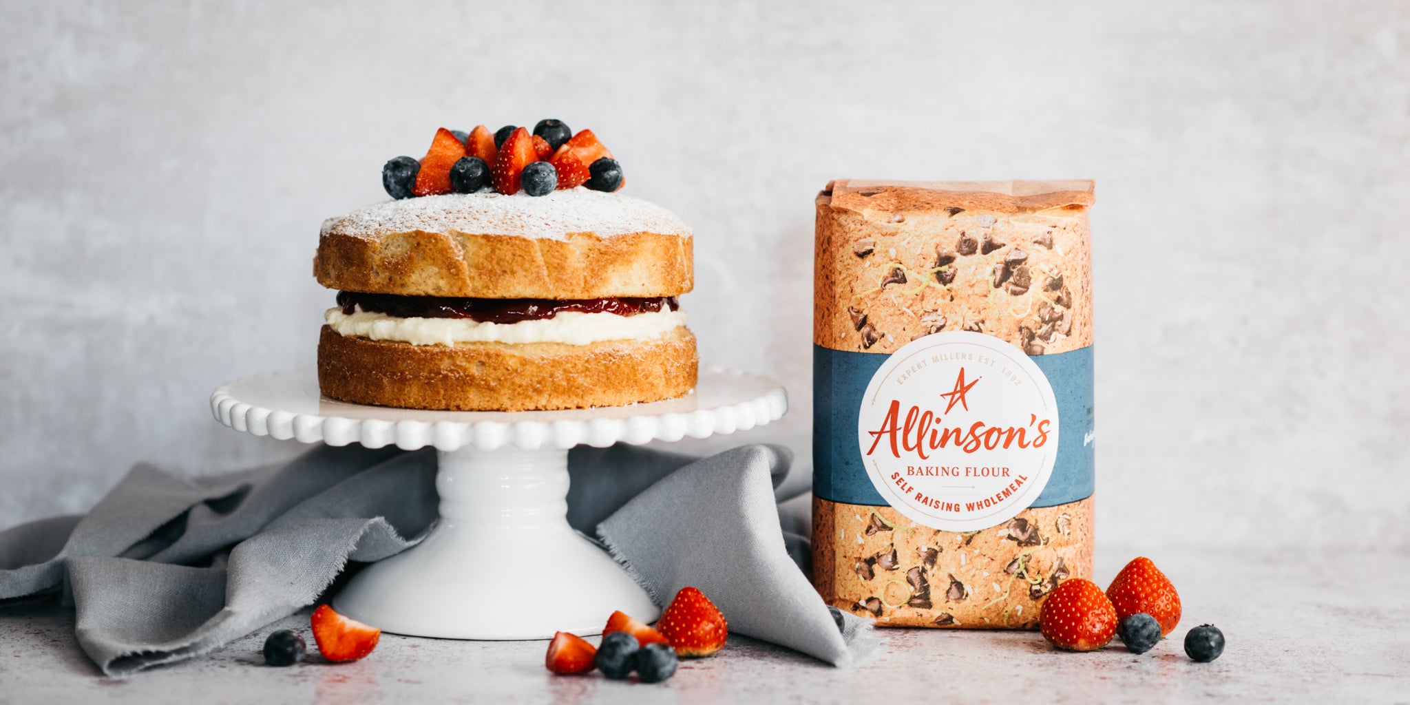 Wholemeal Victoria Sponge on a cake stand, next to a bag of Allinson's Wholemeal Flour and scattered with fresh summer fruits