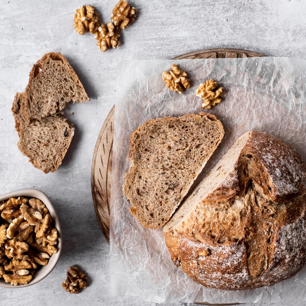 A loaf of chunky walnut bread with 2 slices cut and laid flat on a round wooden board. Some walnuts are scattered in front of and next to the loaf. The image is shot from above.