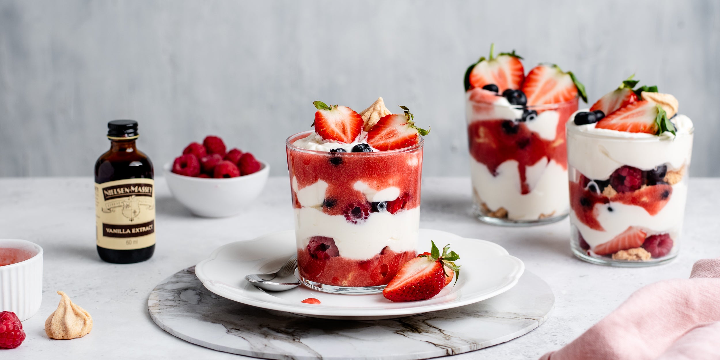 Summer Berry Eton Mess served in glasses, next to a bowl of fresh raspberries and a bottle of Nielsen-Massey Vanilla extract