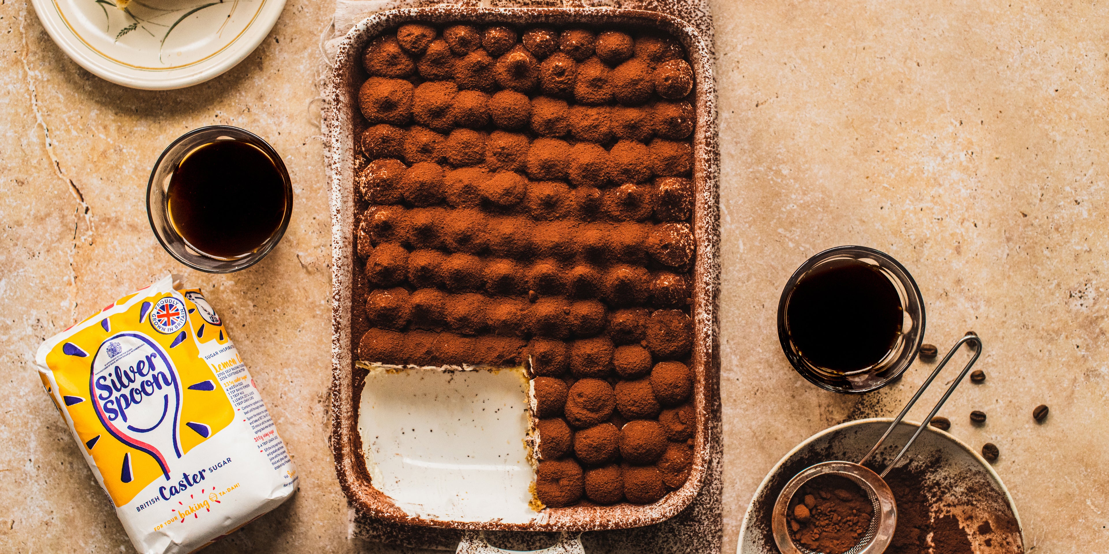 Vegan Tiramisu top view, with a generous slice cut out of it, served next to a bag of Silver Spoon caster sugar, coffee and cocoa powder