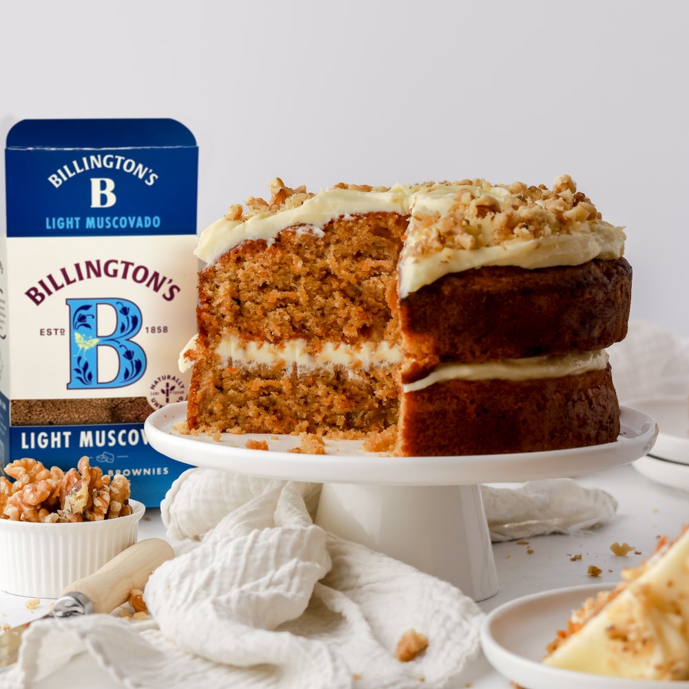 A box of Billington's Light Muscovado Sugar next to a carrot cake topped with buttercream and walnuts which has a large slice cut out of it.
